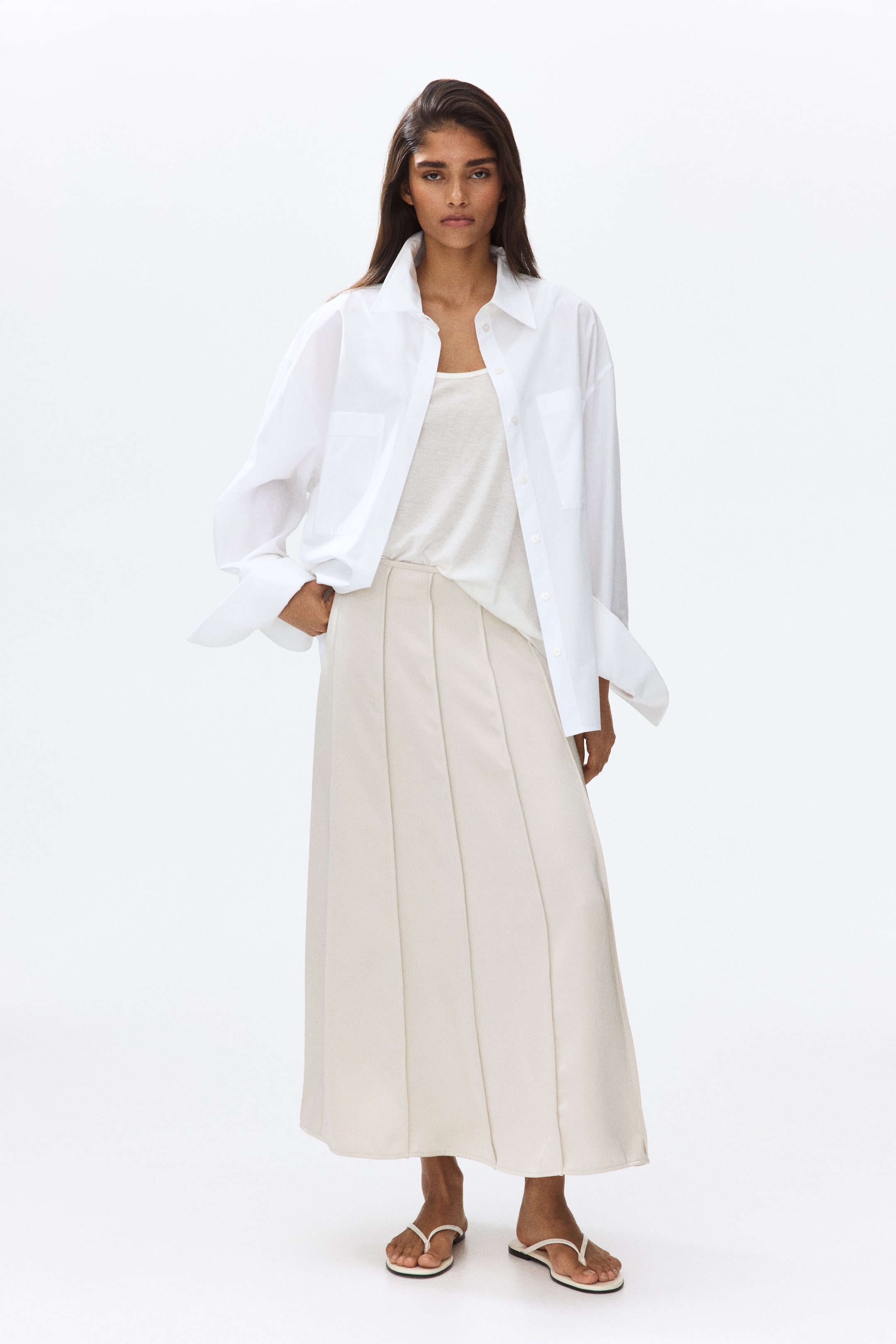 View larger image: The model is wearing a flared maxi skirt crafted from softly draping satin, highlighted by sewn-in pleats down the front. A light tank top is neatly tucked into the skirt, complemented by a white shirt layered over the waist.