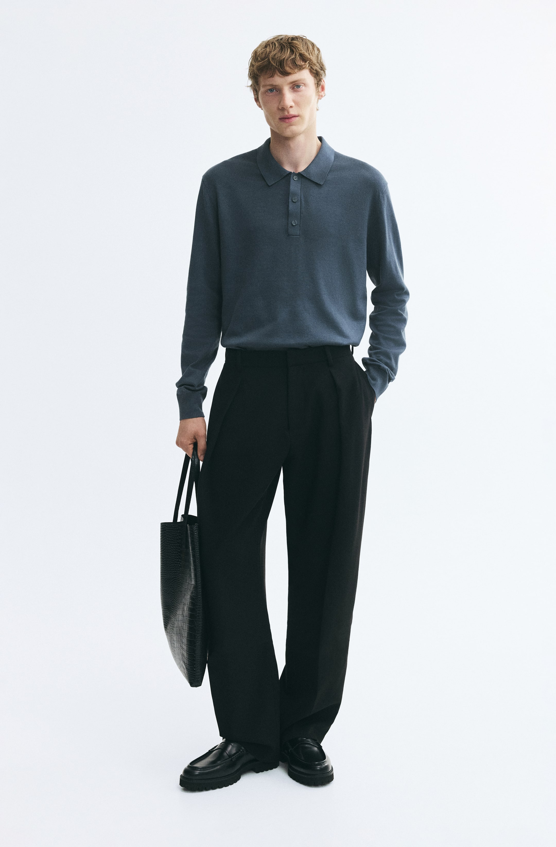 Visualizza l'immagine ingrandita: Il modello indossa una polo a maniche lunghe in maglia fine, blu-grigio, infilata in pantaloni neri a gamba larga. La maglia presenta un colletto classico, una chiusura con bottoni e polsini a costine.