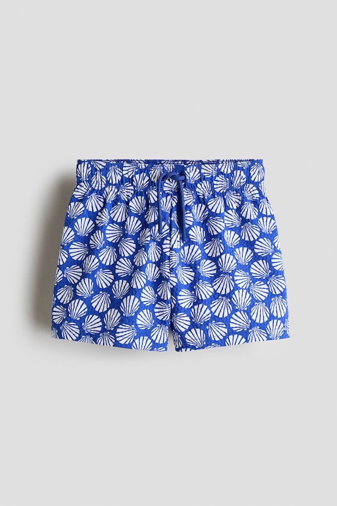 Shorts are displaying a blue and white seashell pattern on a textured fabric. They are featuring an elasticated waistband with a blue drawstring tied at the front. - 1
