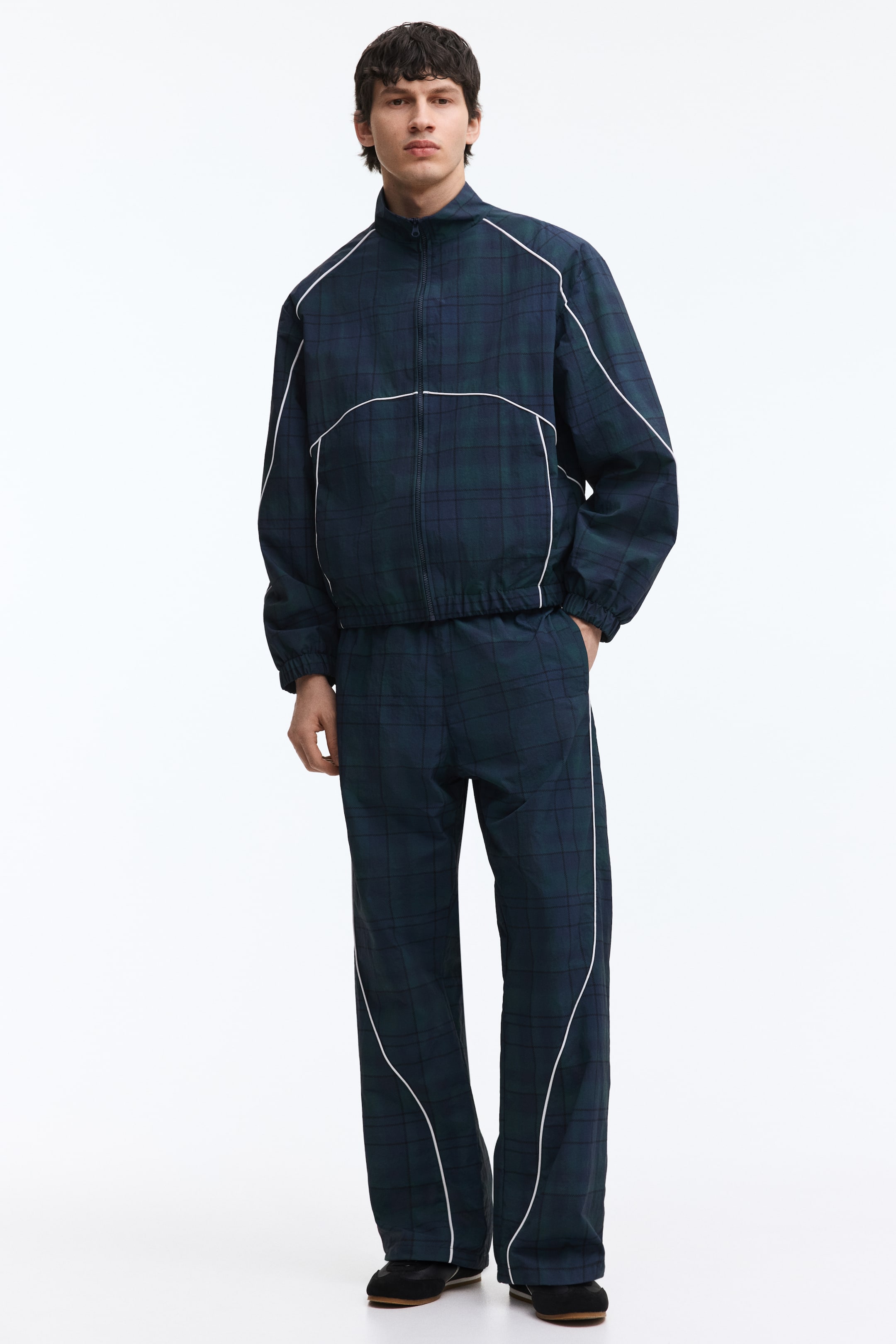 View larger image: A dark green and blue plaid tracksuit featuring a full-zip jacket with a stand-up collar, elasticated cuffs, and an elasticated hem, along with matching straight-leg pants. White piping outlines the jacket's shoulders, sleeves, and a curved chest design, while also running down the outer sides of the pants with a curved detail.