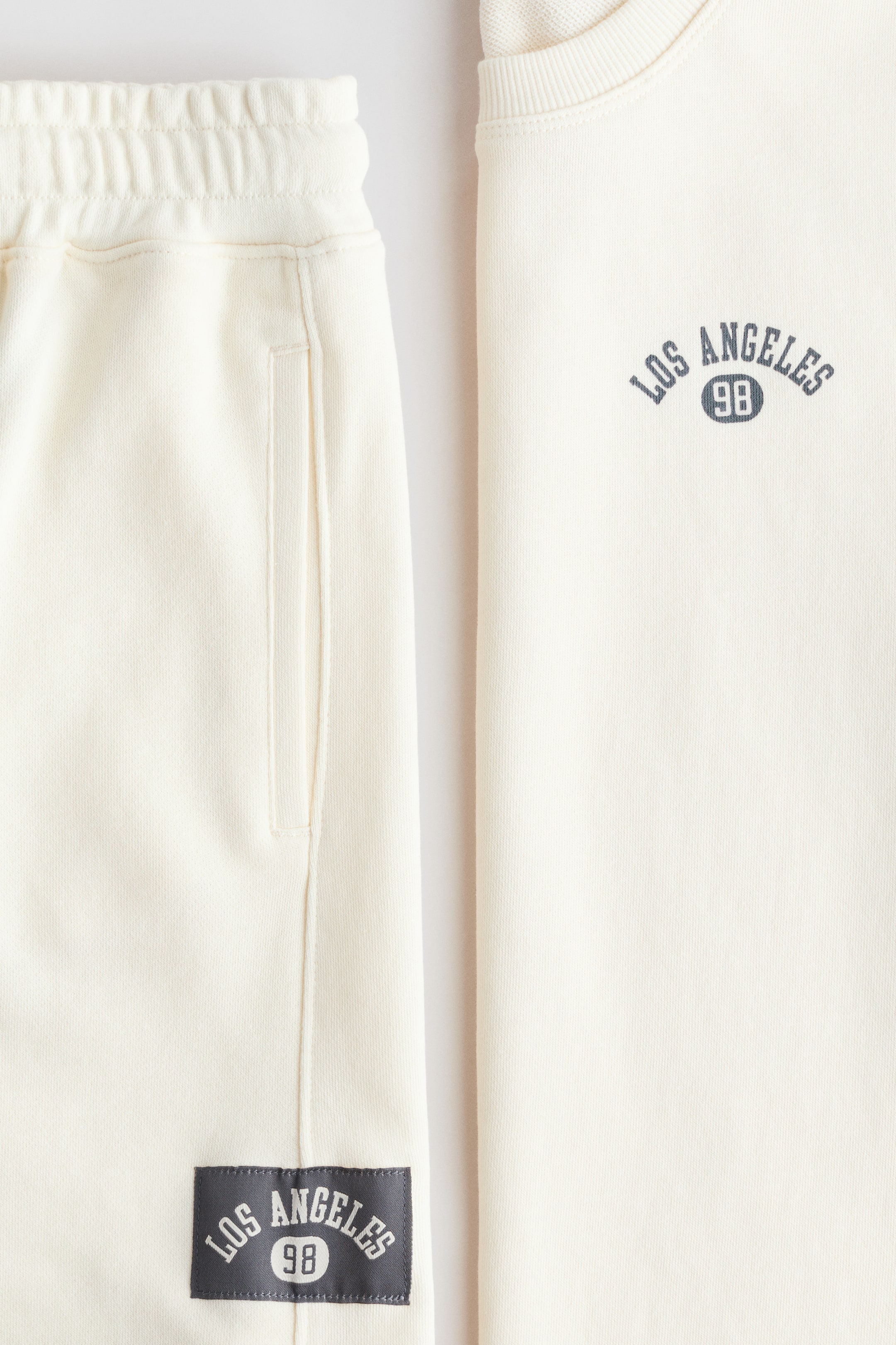 Ver imagen más grande: Un conjunto de ropa de descanso en color crema que incluye una sudadera y pantalones de chándal a juego. La sudadera presenta un cuello redondo acanalado y un gráfico "Los Ángeles 98" en el pecho, mientras que los pantalones de chándal tienen una cintura elástica, un bolsillo lateral y una etiqueta tejida "Los Ángeles 98" en la parte inferior de la pierna.