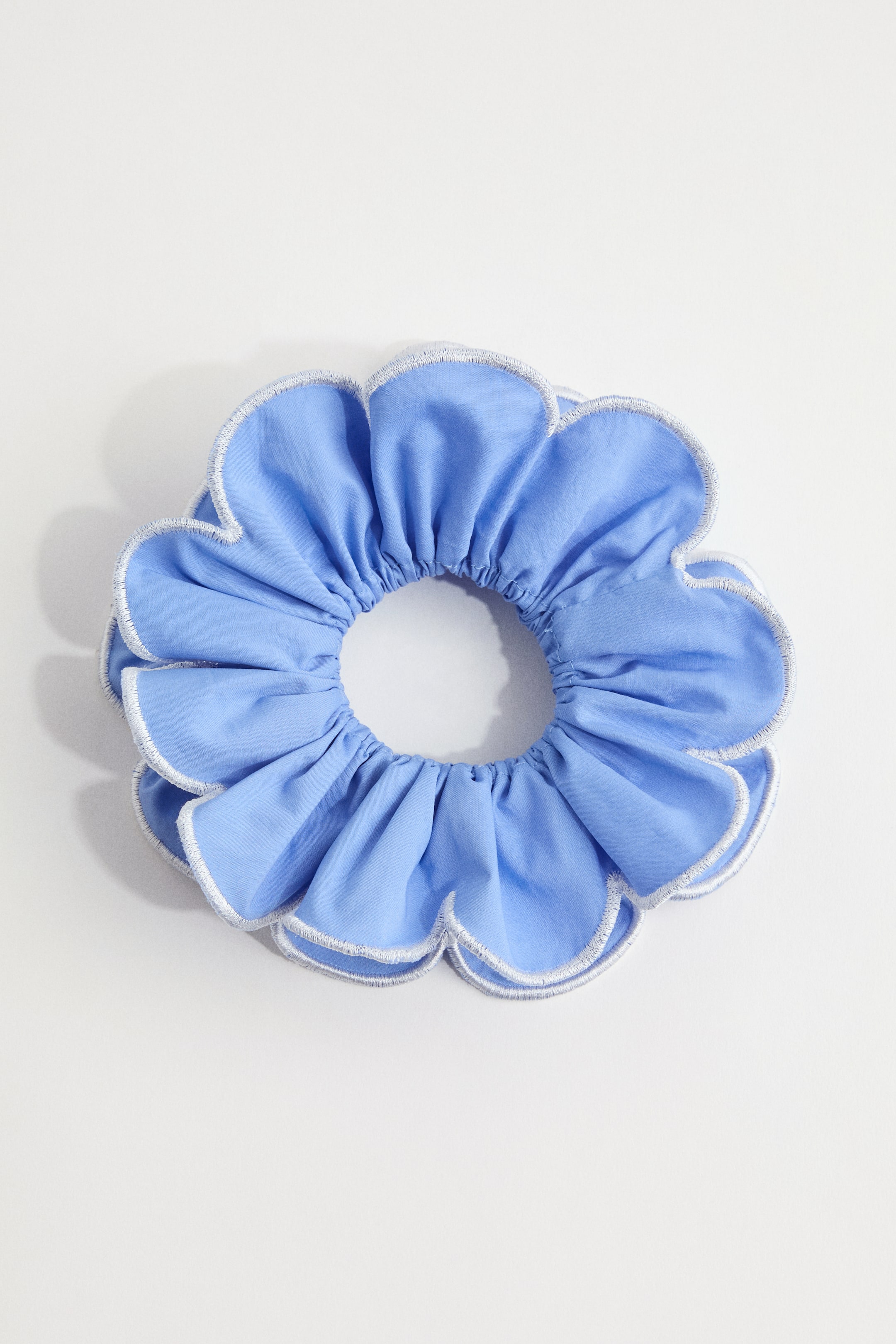 Agrandir l'image: Un chouchou est présenté en vue de dessus, fabriqué à partir d'un tissu bleu clair. Il est façonné comme une fleur avec plusieurs pétales festonnés, chacun bordé de coutures blanches, et le tissu central est froncé autour d'un élastique.