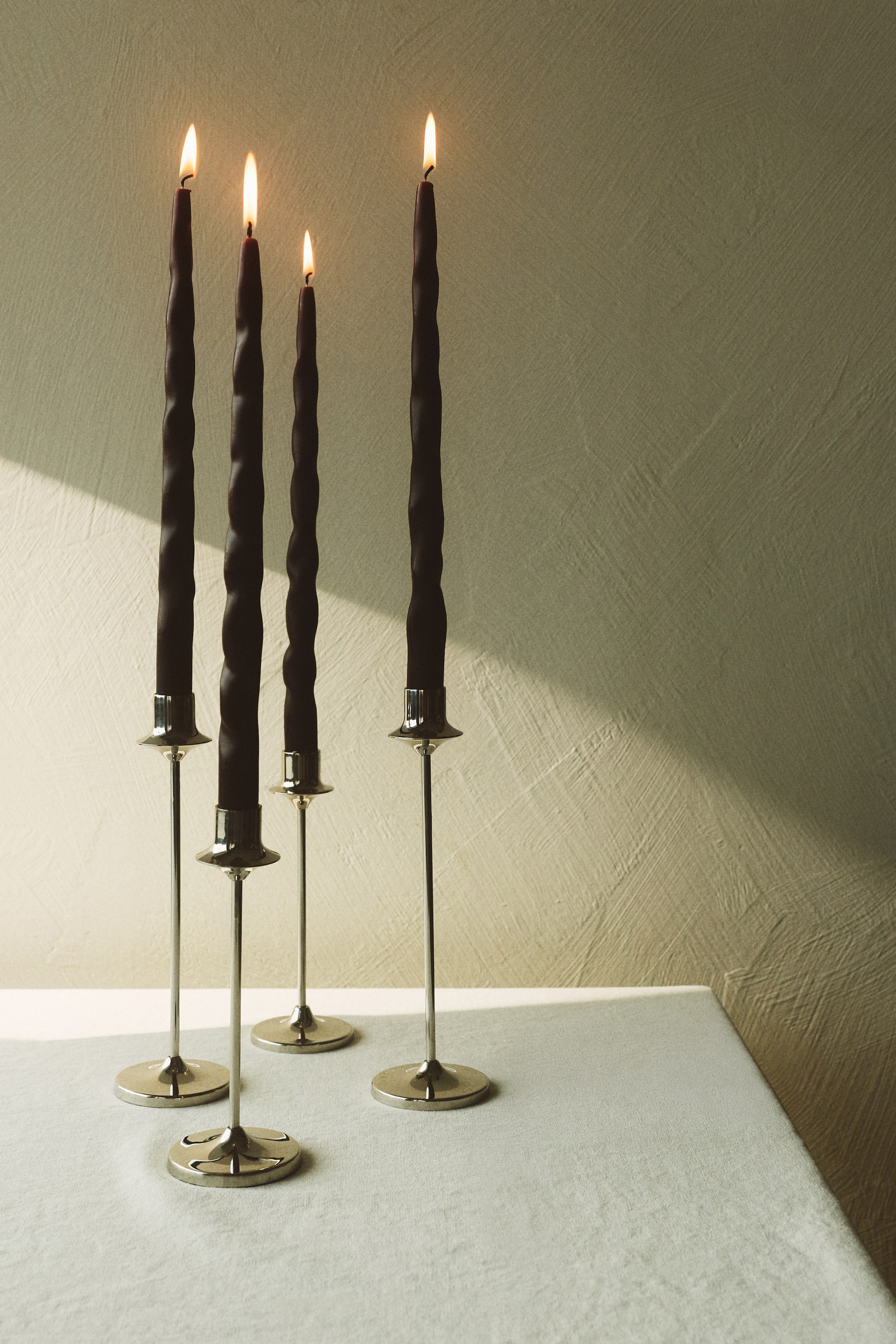 Se større billede: Fire mørke, tilspidsede stearinlys, hver med et håndformet bølget design, står oprejst i slanke, metalliske lysestager i forskudte højder. Deres væger brænder og producerer flammer.