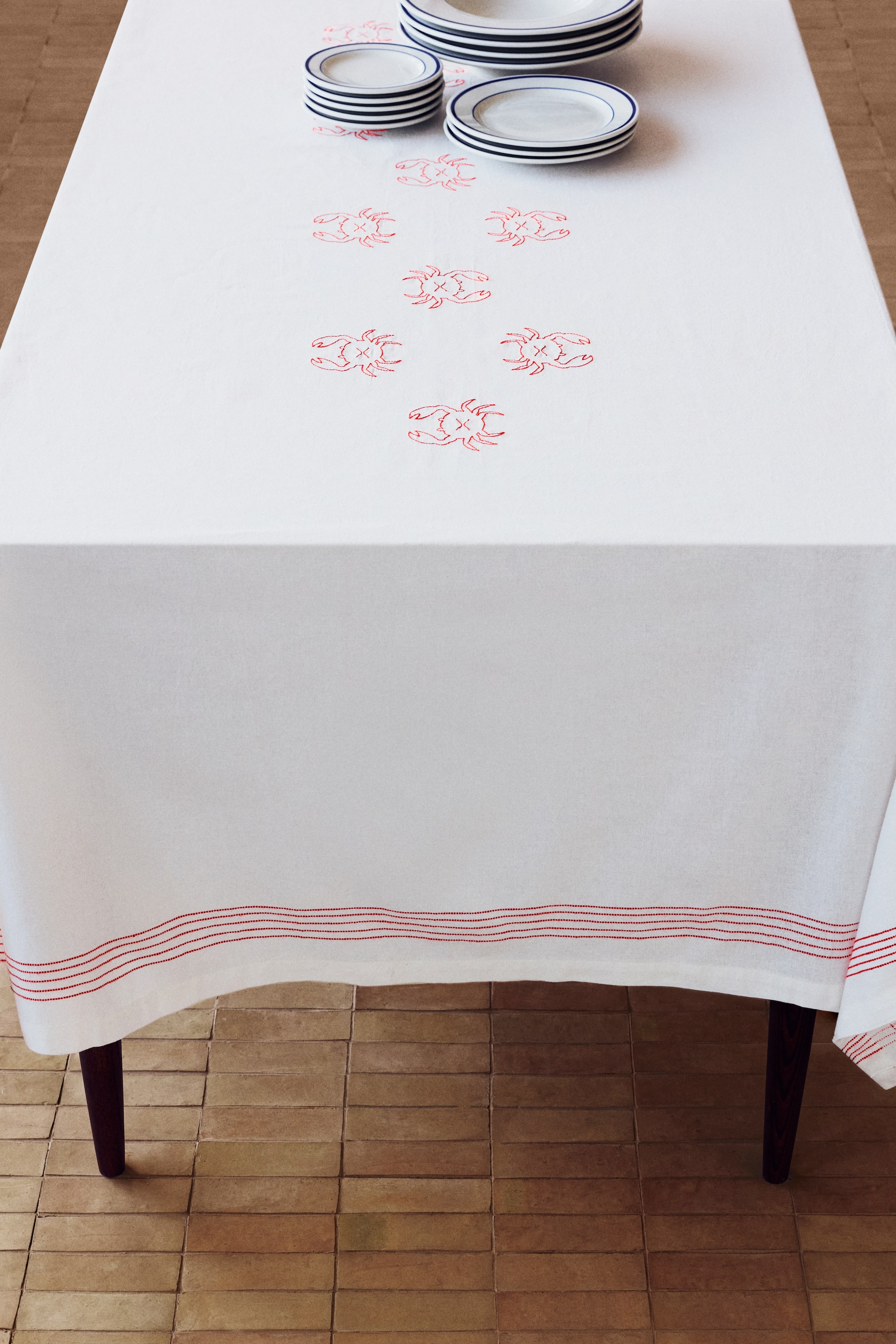 Voir une image plus grande: Une nappe blanche recouvre une table, arborant des motifs de crabe rouge brodés sur sa surface supérieure et des rayures rouges cousues le long de son bord inférieur drapé. Des assiettes blanches empilées avec des rebords sombres reposent sur la nappe.
