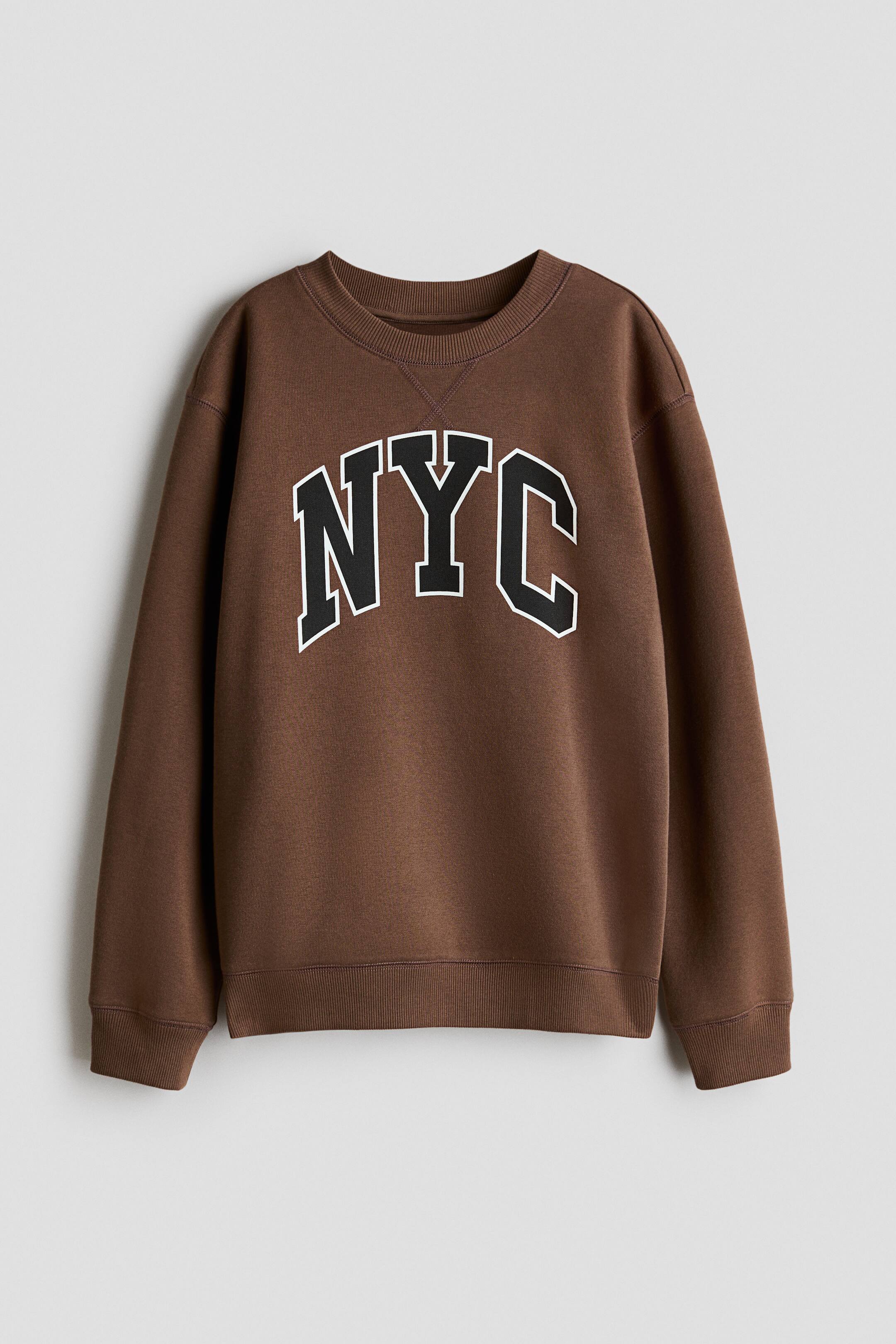 Büyük resmi görüntüle: Göğüs kısmında beyaz konturlu, siyah blok harflerle "NYC" yazısı bulunan kahverengi bir sweatshirt. Ayrıca nervürlü bir bisiklet yaka, nervürlü manşetler ve nervürlü bir etek ucu ile öne çıkıyor.