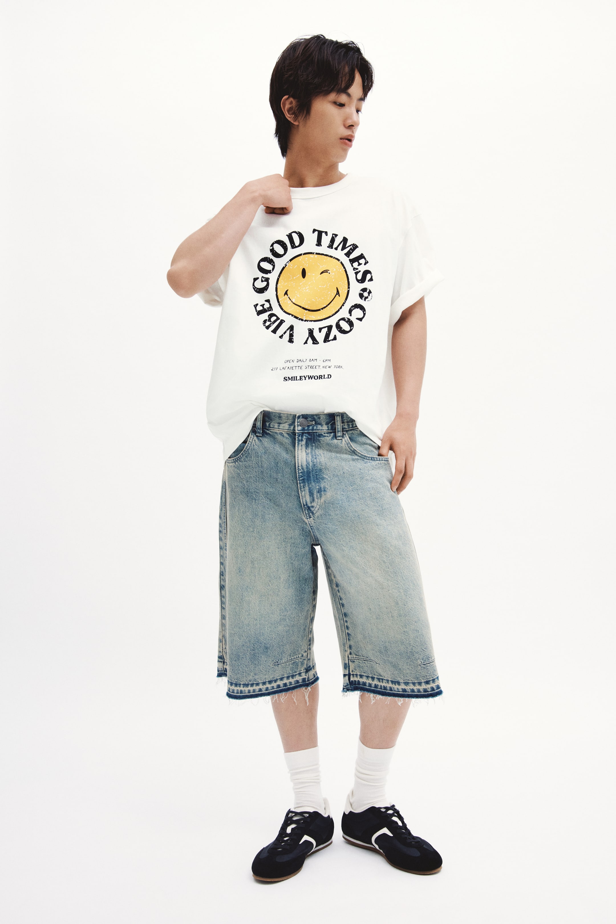 拡大画像を表示: 白のオーバーサイズTシャツは、フロントに大きな円形グラフィックを特徴としており、黄色のウィンクするスマイリーフェイスが、傷んだ黒いテキストで囲まれています。「GOOD TIMES & COZY VIBE」と「SMILEYWORLD」が下部に表示されています。このTシャツは、ショートカフススリーブとクルーネックを備えています。