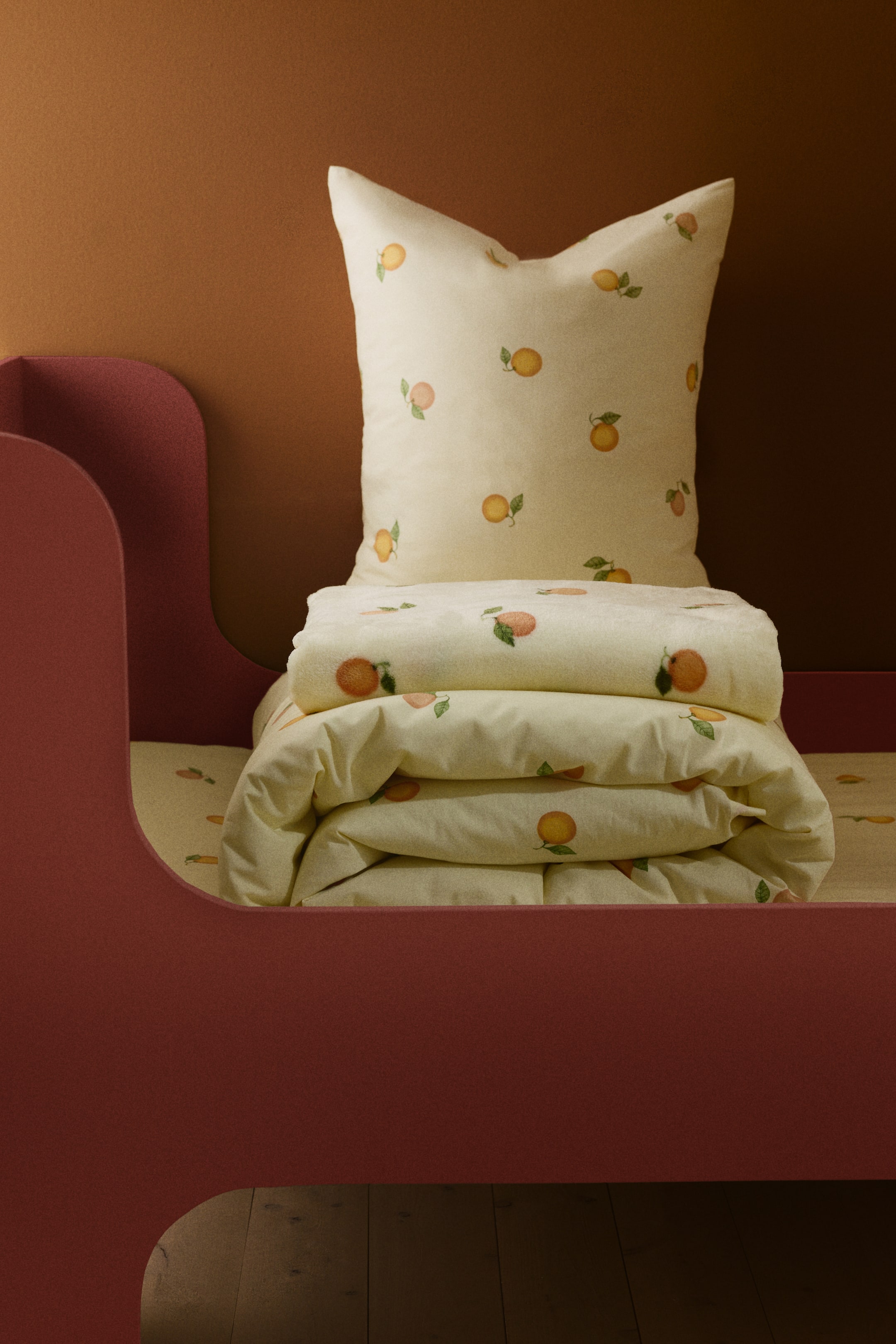 Nagyobb kép megtekintése: "Egy paplanhuzat garnitúra, amely világos sárga anyagból készült, teljes felületén narancsokkal és zöld levelekkel díszítve. A képen egy párnahuzat áll függőlegesen, szemből látható, és egy gondosan összehajtott paplanhuzat."