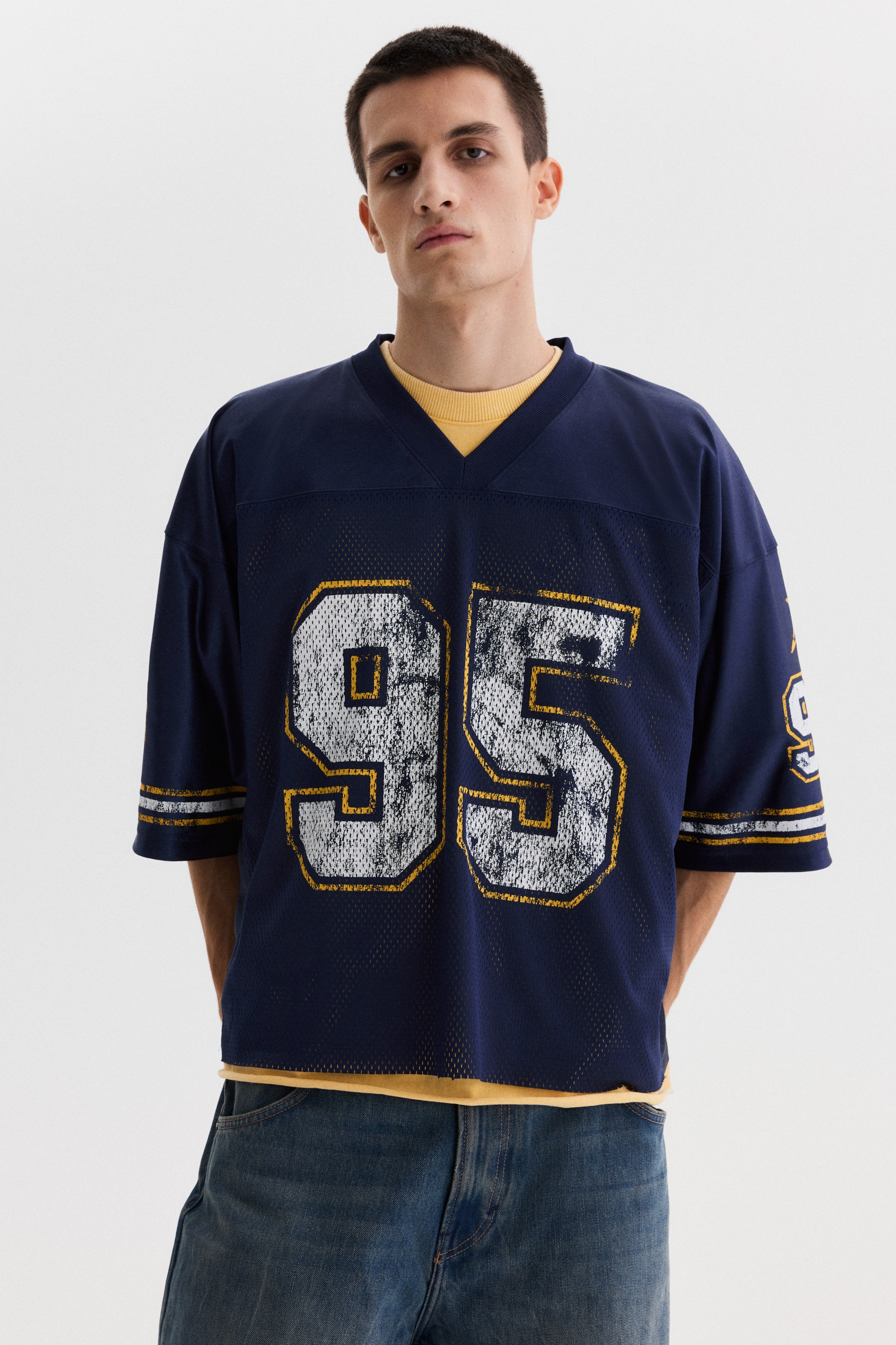 View larger image: A navy-blue mesh jersey showcases a V-neck, short, wide sleeves with gold and white stripes, and large, distressed silver-white numbers "95" with a gold outline on the front. A yellow T-shirt is layered underneath.