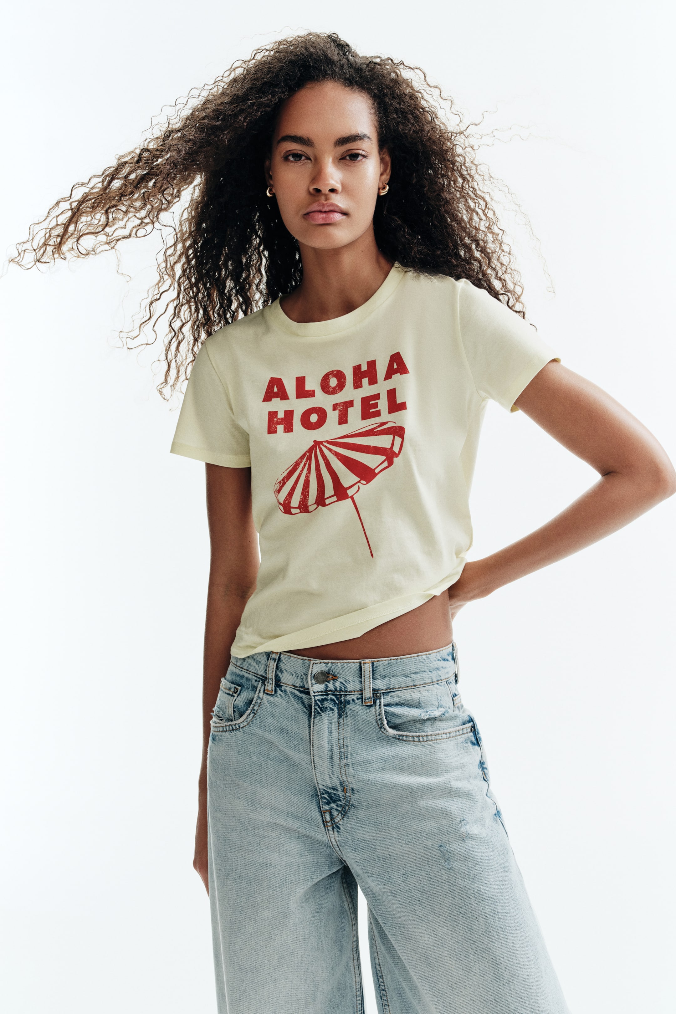 View larger image: A light yellow t-shirt menampilkan grafik "ALOHA HOTEL" merah yang kelihatan usang di atas payung pantai berjalur merah dan putih yang juga kelihatan usang. Ia dipakai dengan lengan pendek, leher bulat, dan potongan yang sedikit pendek.