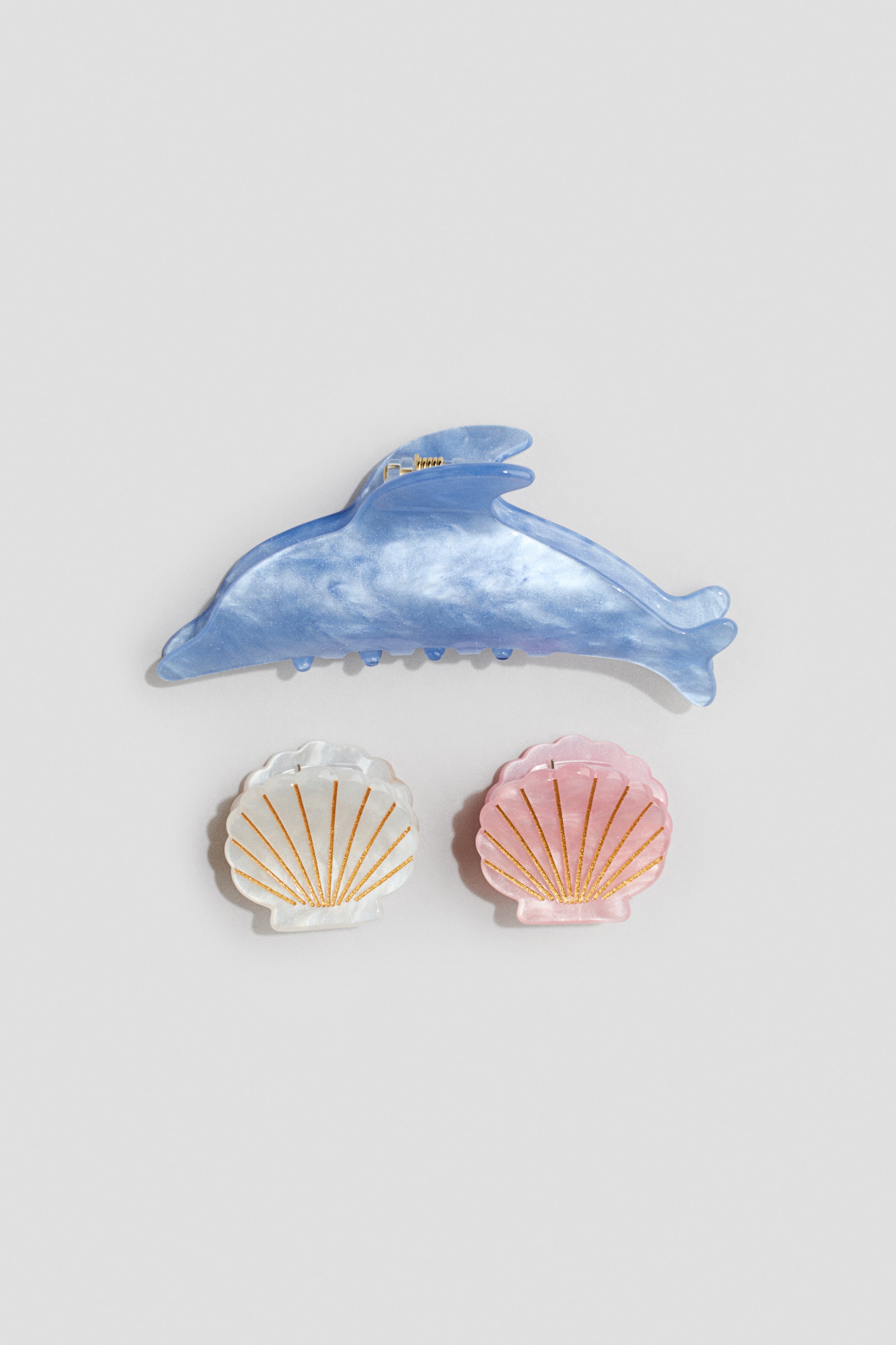 Agrandir l'image: Les pinces à cheveux se présentent sous forme d'un ensemble de trois en plastique marbré et scintillant. Une pince en forme de dauphin bleu est positionnée au-dessus de deux pinces en forme de coquillage, qui apparaissent en blanc et rose avec des lignes dorées rayonnantes.