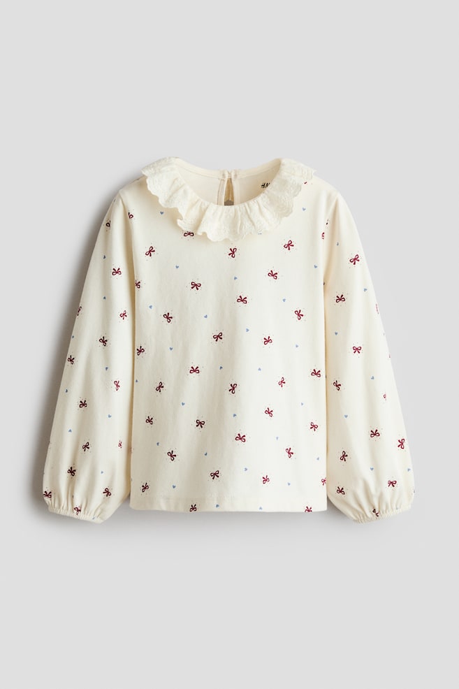A cream-coloured long-sleeved top is shown from the front, featuring an all-over pattern of small red bows and tiny blue hearts. The garment features a ruffled, eyelet-trimmed collar and long sleeves ending in gathered, elasticated cuffs. - 1