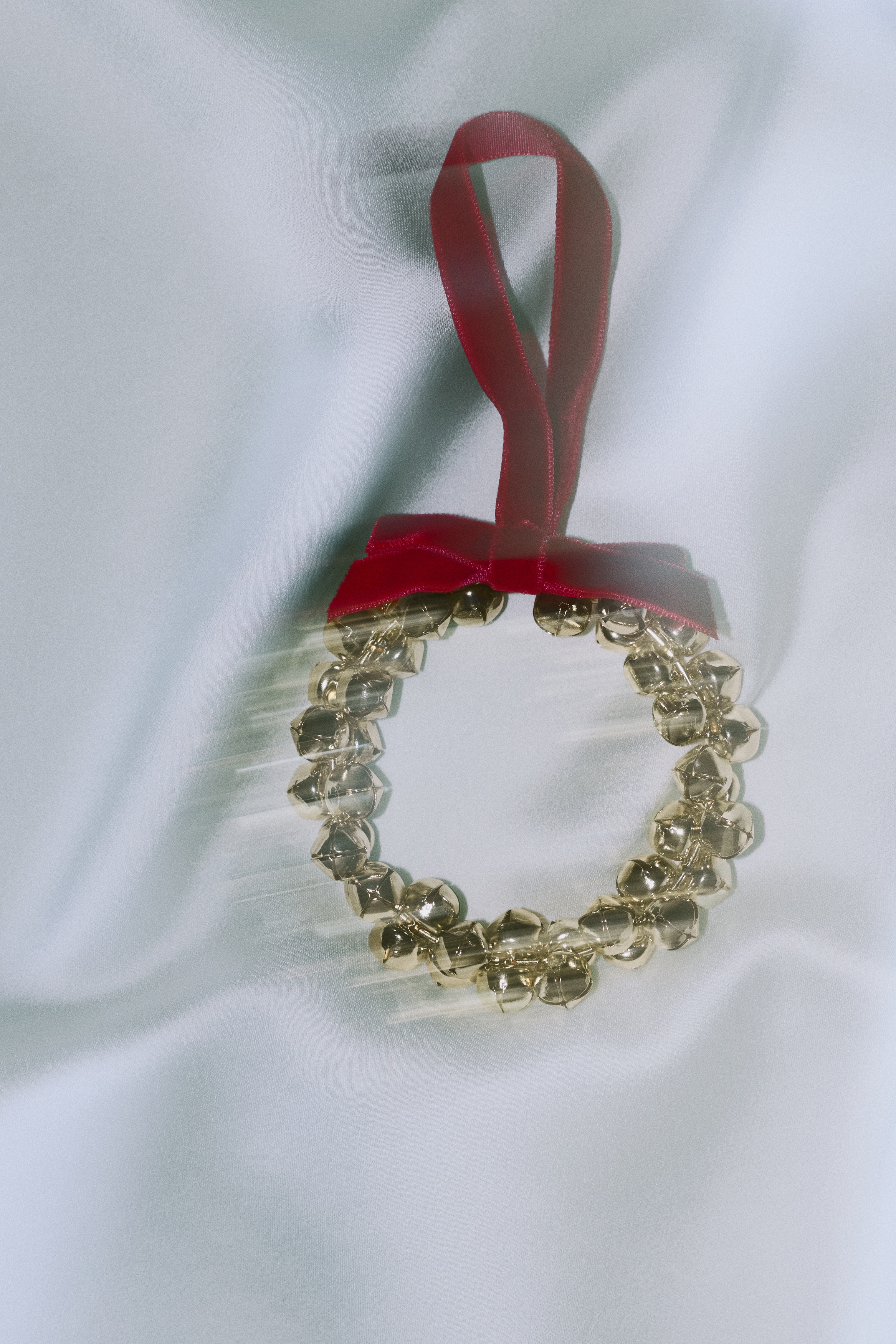Una vista FRONTAL de una corona circular, compuesta por numerosos cascabeles de color dorado, cuelga de una cinta de terciopelo rojo. La cinta está adornada con un pequeño lazo donde se conecta a la parte superior de la corona.