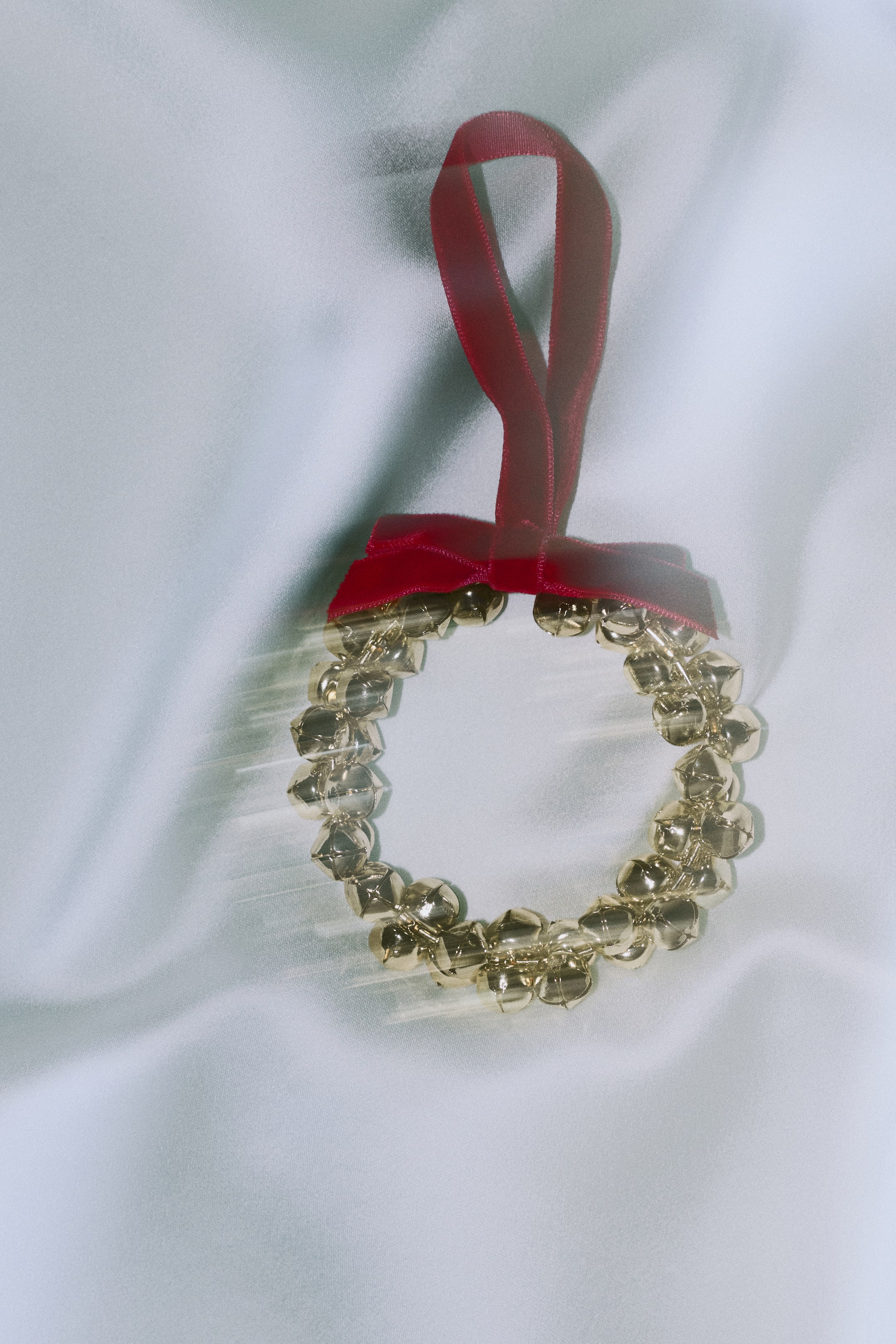 View larger image: A circular wreath ornament composed of shiny, gold-toned jingle bells, featuring a red velvet ribbon tied into a bow at the top with an attached hanging loop. The ornament is presented in a front-facing view, highlighting its textured metallic structure and soft ribbon details.
