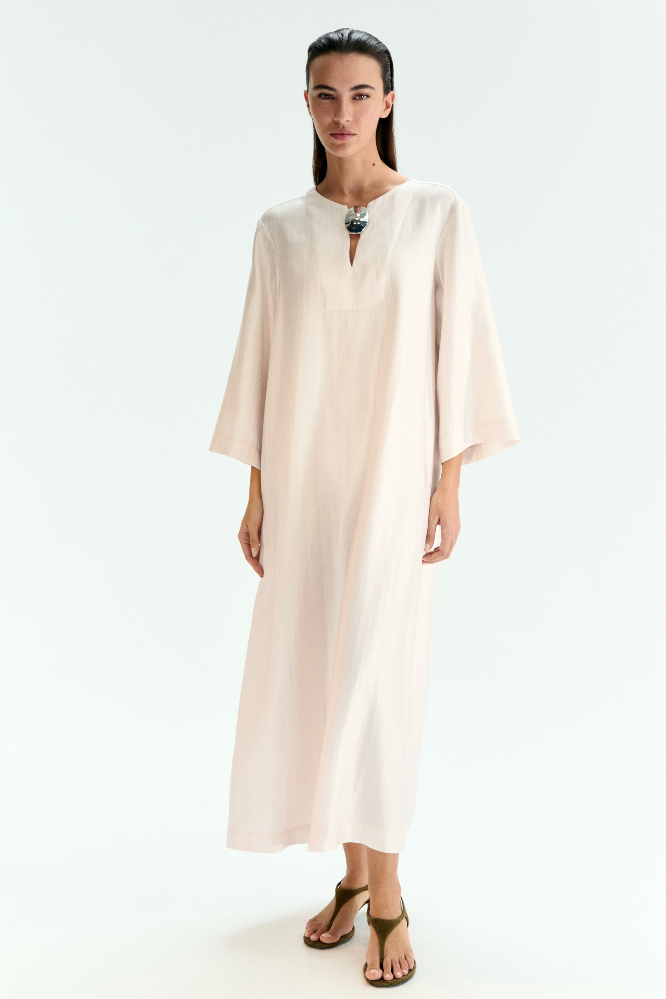 View larger image: The pale pink maxi dress is presenting with a loose, flowing silhouette, facing front. It is featuring a round neckline with a V-split, which is secured by a decorative metallic clasp, and wide, three-quarter length sleeves.