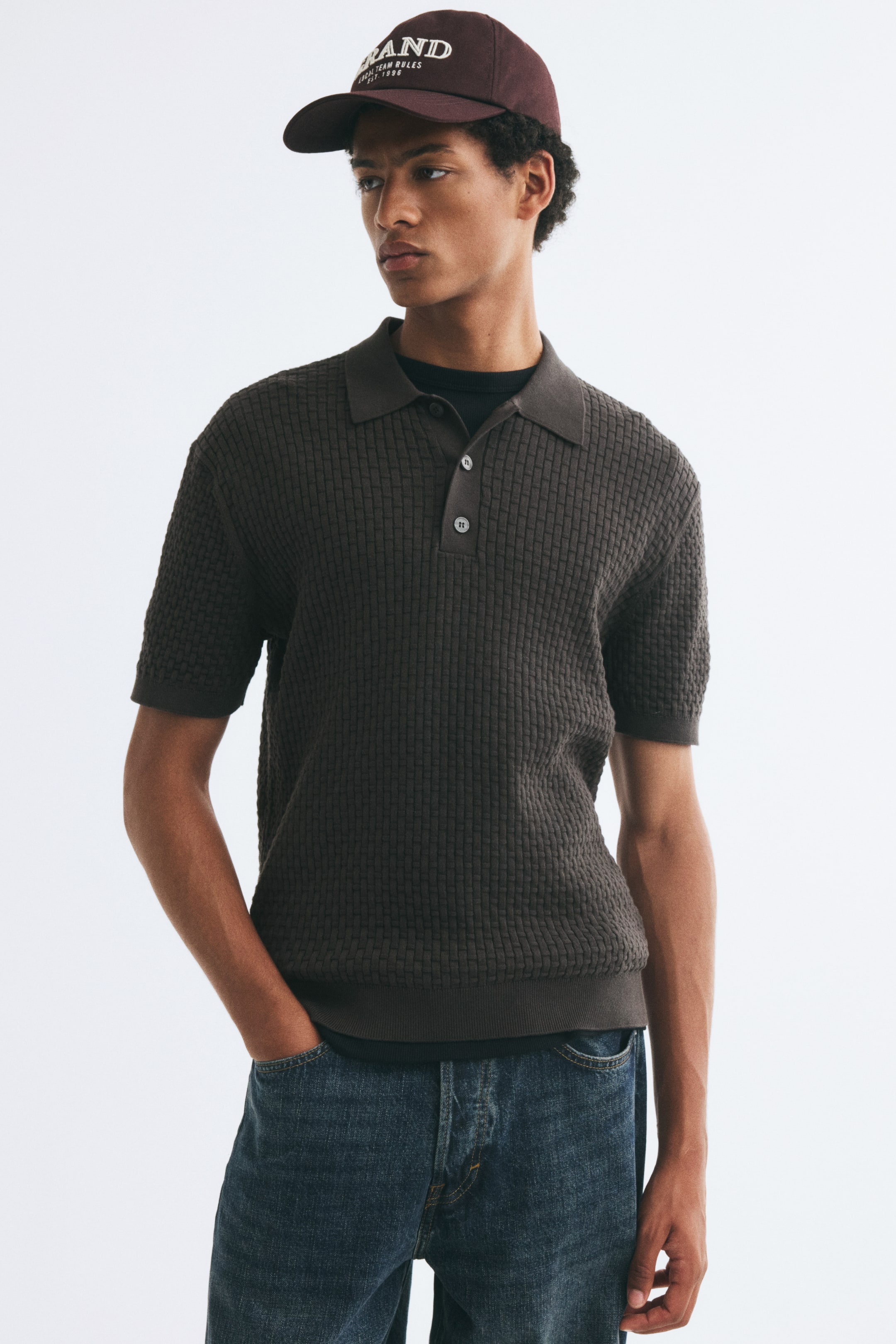 View larger image: The model is wearing a dark brown textured knit polo shirt layered over a black crewneck shirt and paired with blue jeans. The short-sleeve polo features a waffle-stitch pattern, a ribbed collar with a button placket, and ribbed cuffs and hem.