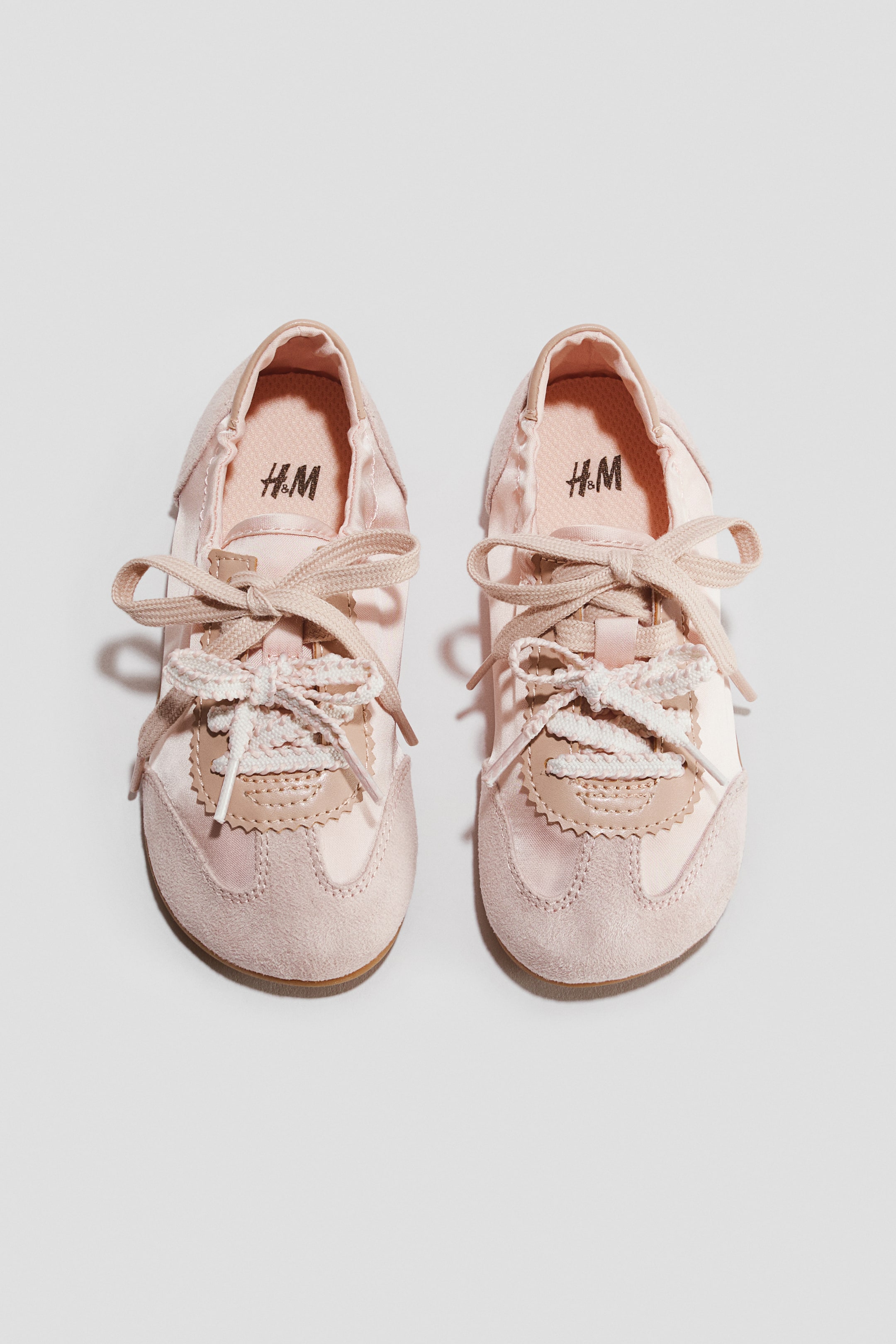 Agrandir l'image: Deux chaussures pour bébé rose pâle sont présentées en vue de dessus. Elles présentent une tige satinée, des empiècements en suédine sur le bout et les côtés, ainsi que des panneaux d'œillets en similicuir beige aux bords festonnés. Des lacets en ruban rose pâle sont noués en nœuds sur des lacets blancs plus fins, et le logo 'H&M' est visible sur les semelles intérieures.