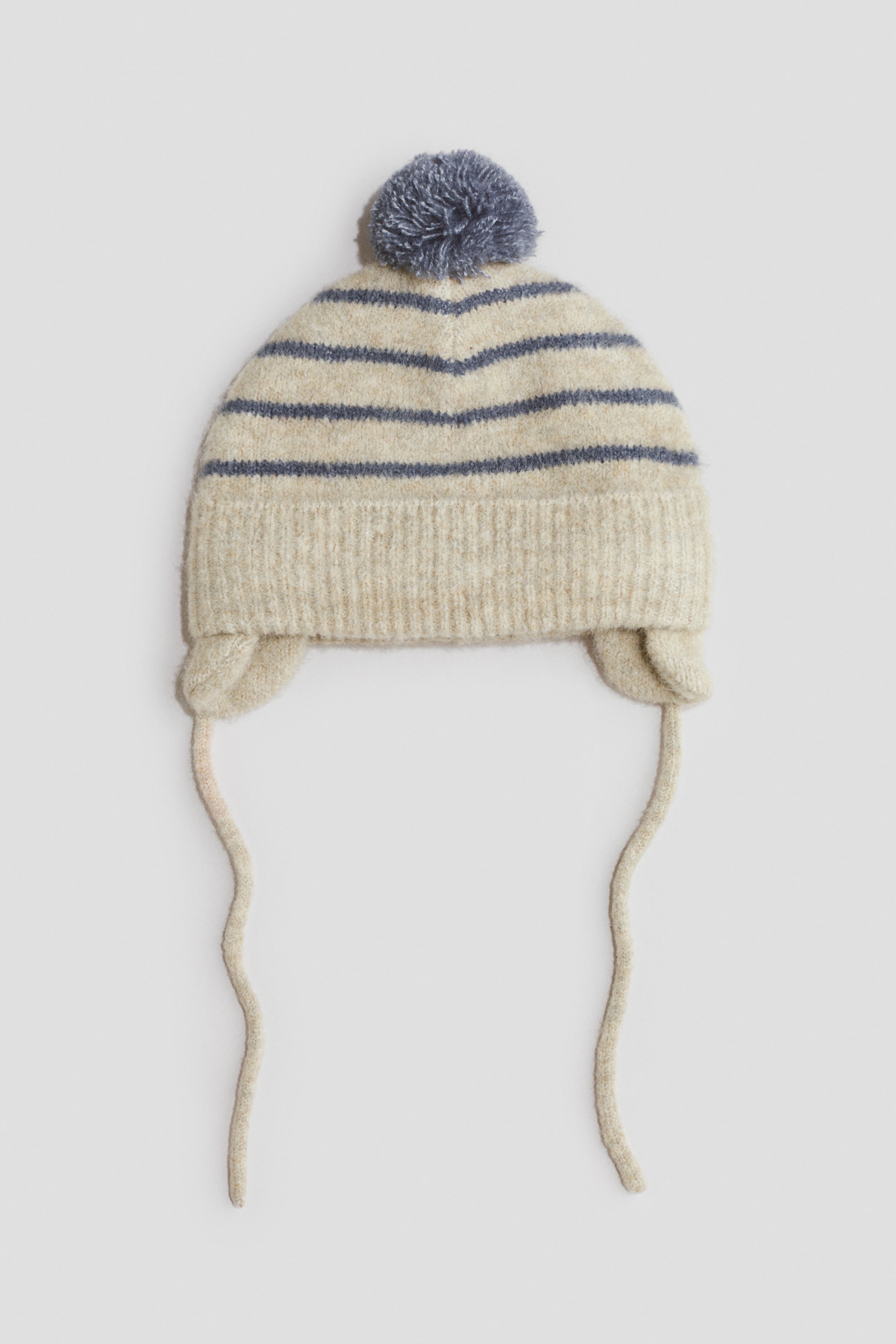 View larger image: A cream knitted hat featuring horizontal blue-grey stripes and a matching pom-pom on top, shown from the front. It is designed with a ribbed brim and ear flaps with long ties.