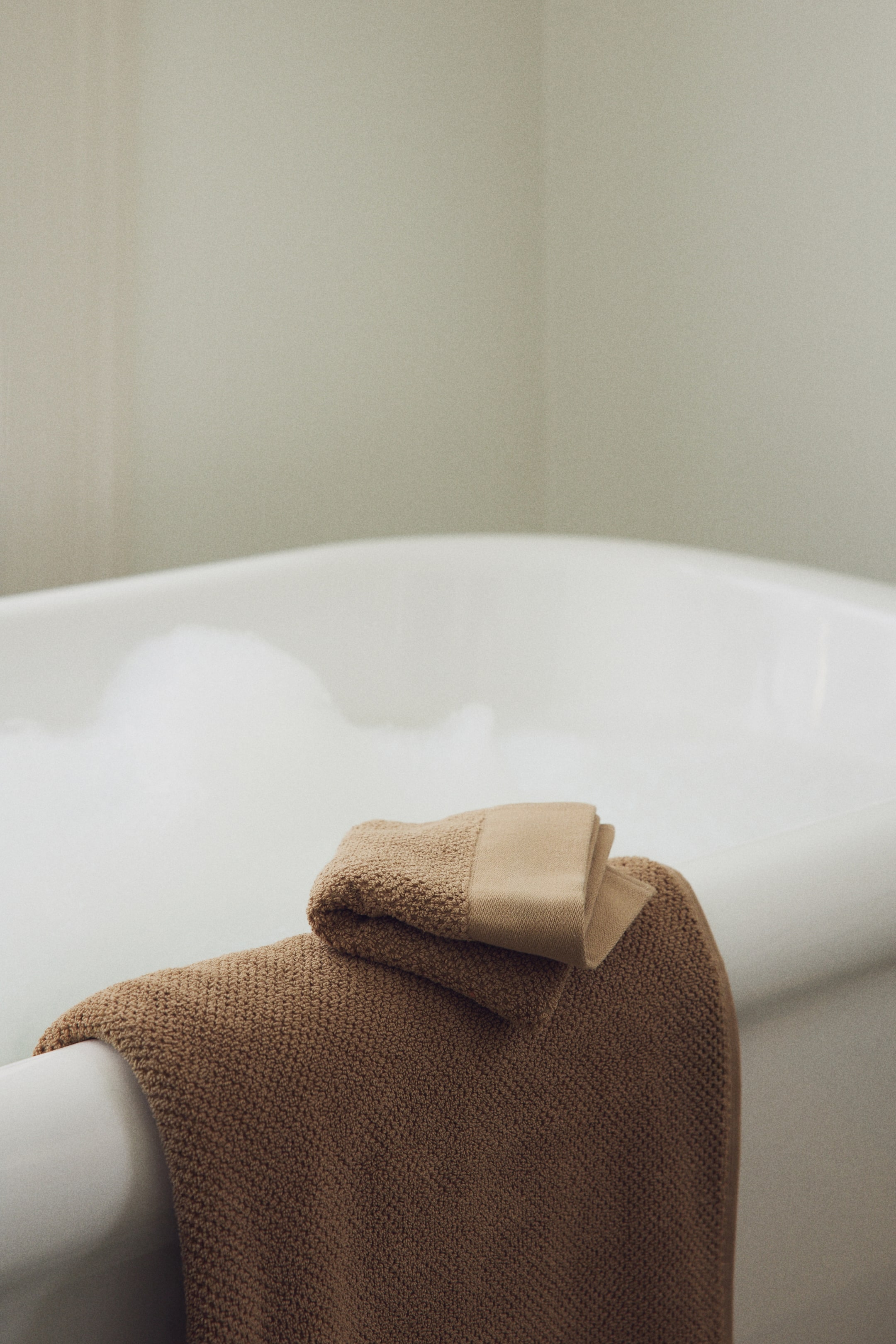 View larger image: A pair of tan terry guest towels is displayed. One towel is folded, showcasing a flat woven hem, and rests on a larger, unfolded terry towel draping over an edge.