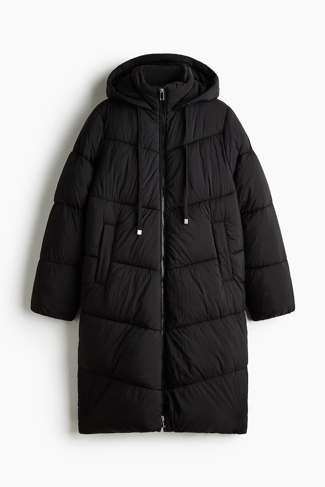 A long black puffer coat is shown from the front, featuring horizontal quilting and a full-length front zip. The coat has an attached hood with adjustable drawstrings and two side welt pockets. - 1