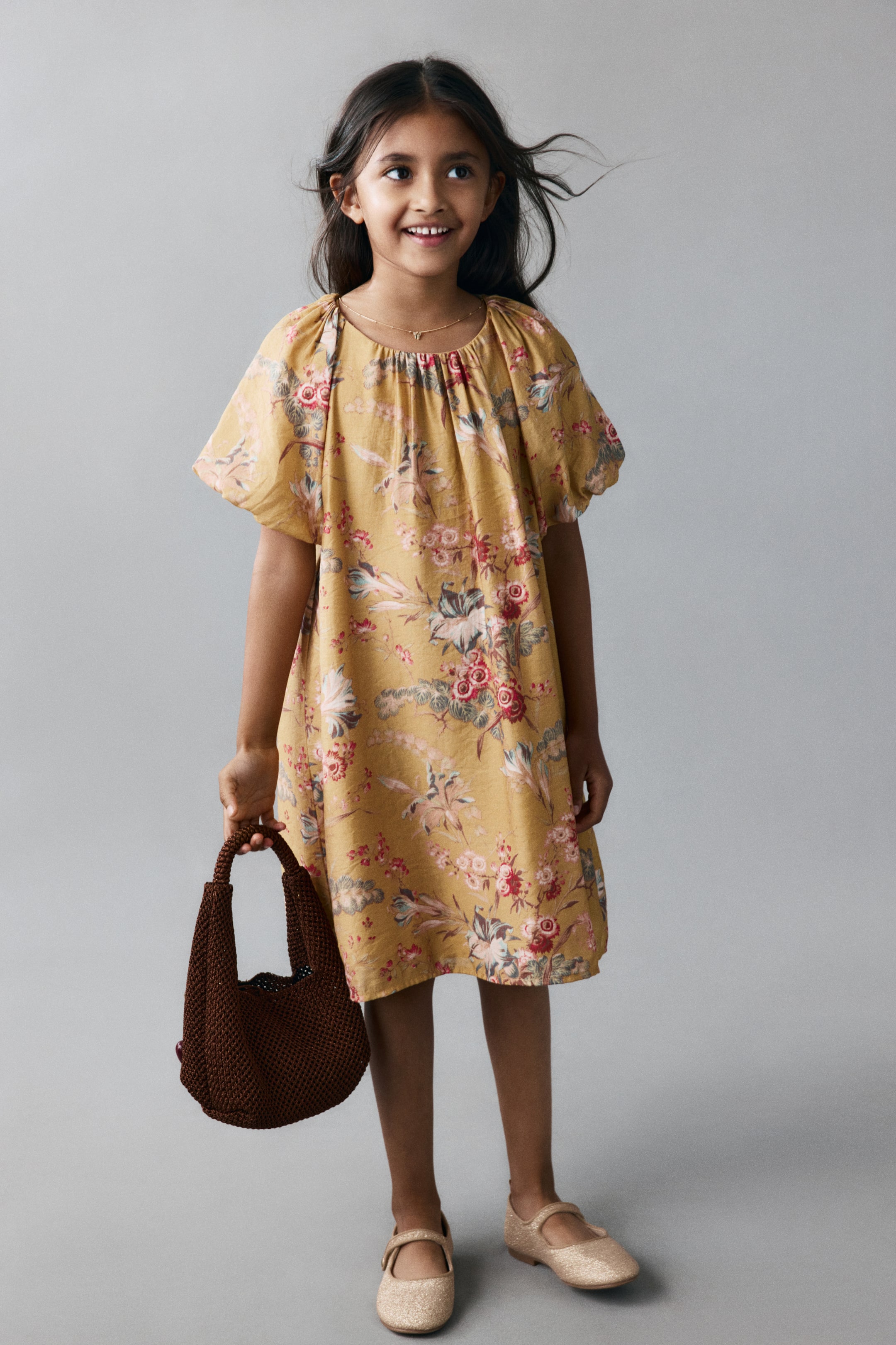 View larger image: A child is wearing a mustard-yellow, floral-patterned A-line dress and is facing the front, showcasing its gathered round neckline and short, voluminous raglan sleeves with elasticated cuffs.