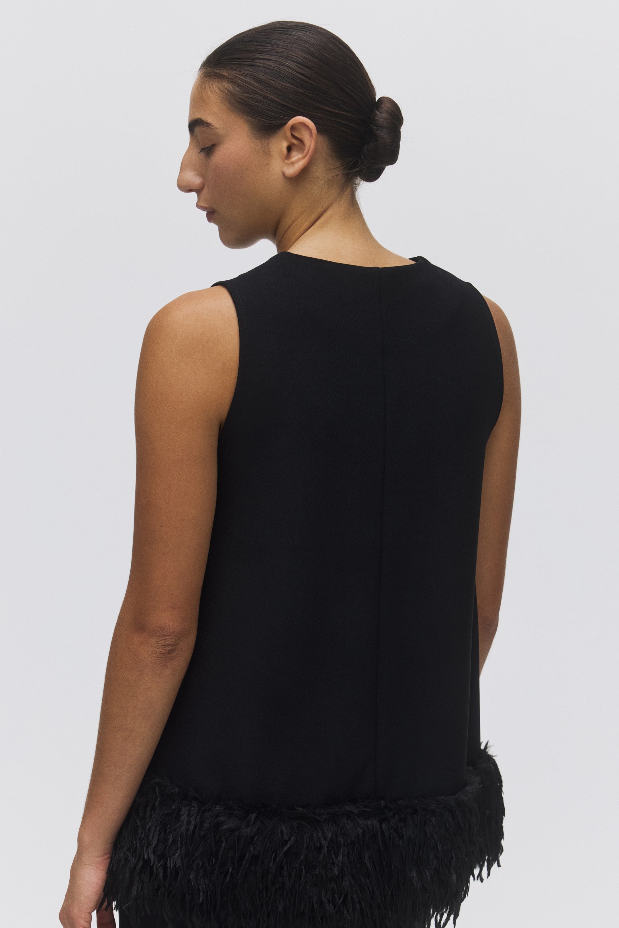 View larger image: A black sleeveless garment is being viewed from the back, featuring a high neckline and a prominent feathered trim along the hem. A vertical seam is running down the center back.