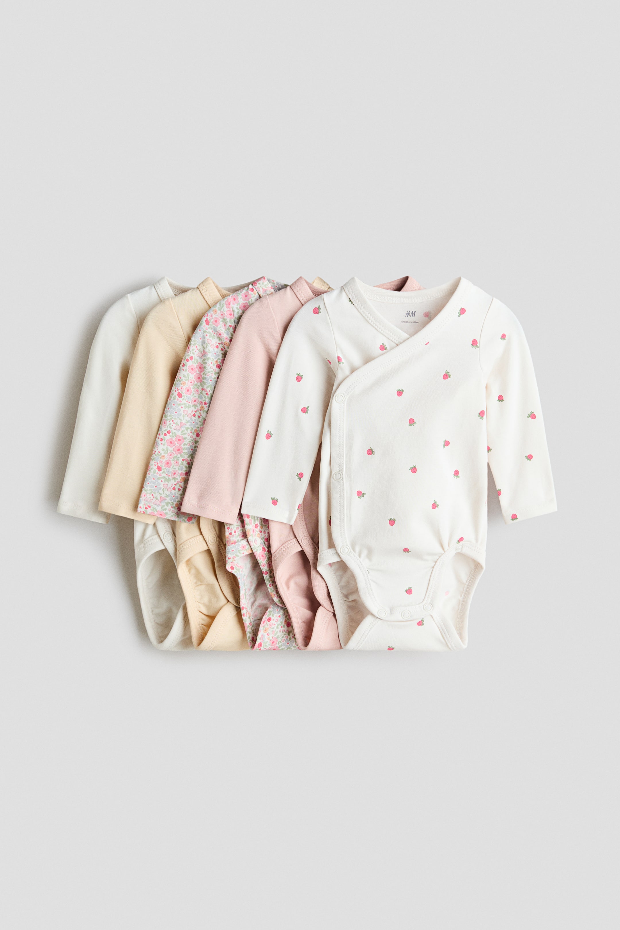 View larger image: Five long-sleeved bodysuits are stacked, facing front. The top bodysuit is cream-coloured with a small strawberry print, featuring a wrapover front with visible press studs along the side and at the bottom, and gathered seams around the leg openings. Other bodysuits are visible behind it in solid light pink, a pink floral print, solid light yellow and solid off-white.
