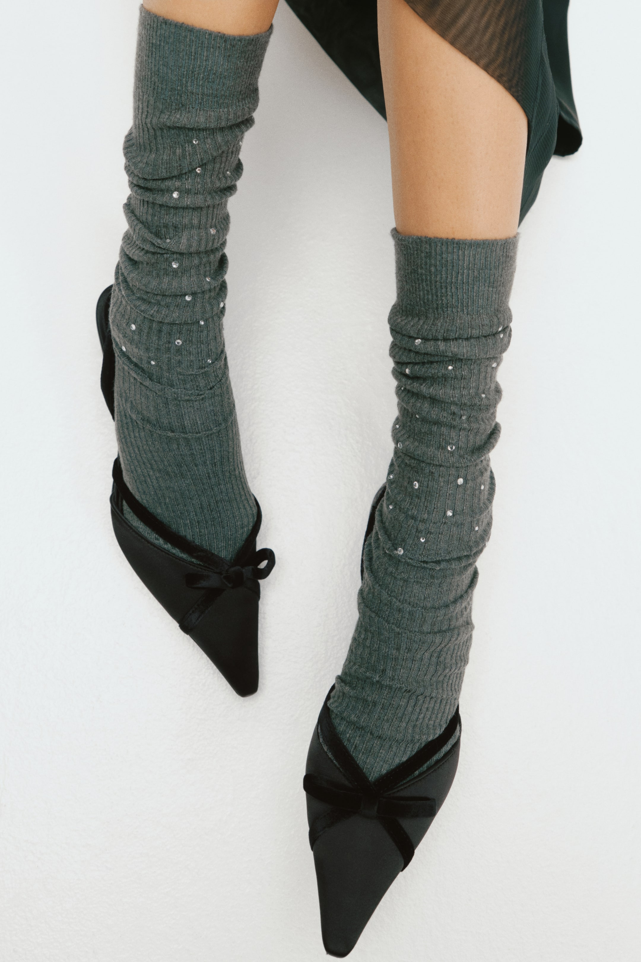 View larger image: Black satin slingback shoes featuring pointed toes and velvet bow details are being worn by the model. The slingbacks are styled over slouchy, grey ribbed socks embellished with small rhinestones.
