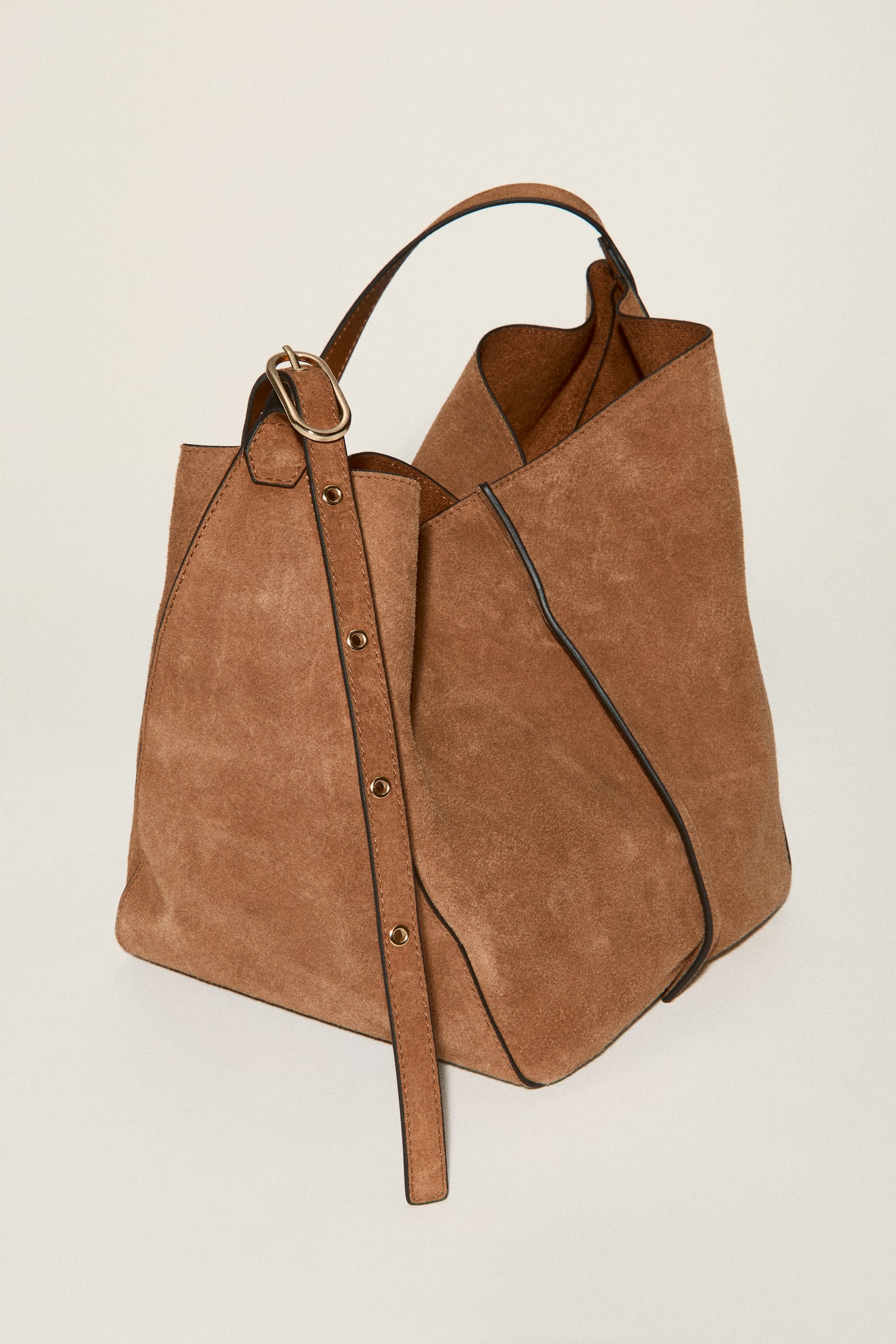 A brown suede shoulder bag is standing upright, showcasing its soft, slouchy silhouette. Dark piping is detailing its seams, and a thin brown strap with an adjustable buckle is extending upwards. - 1