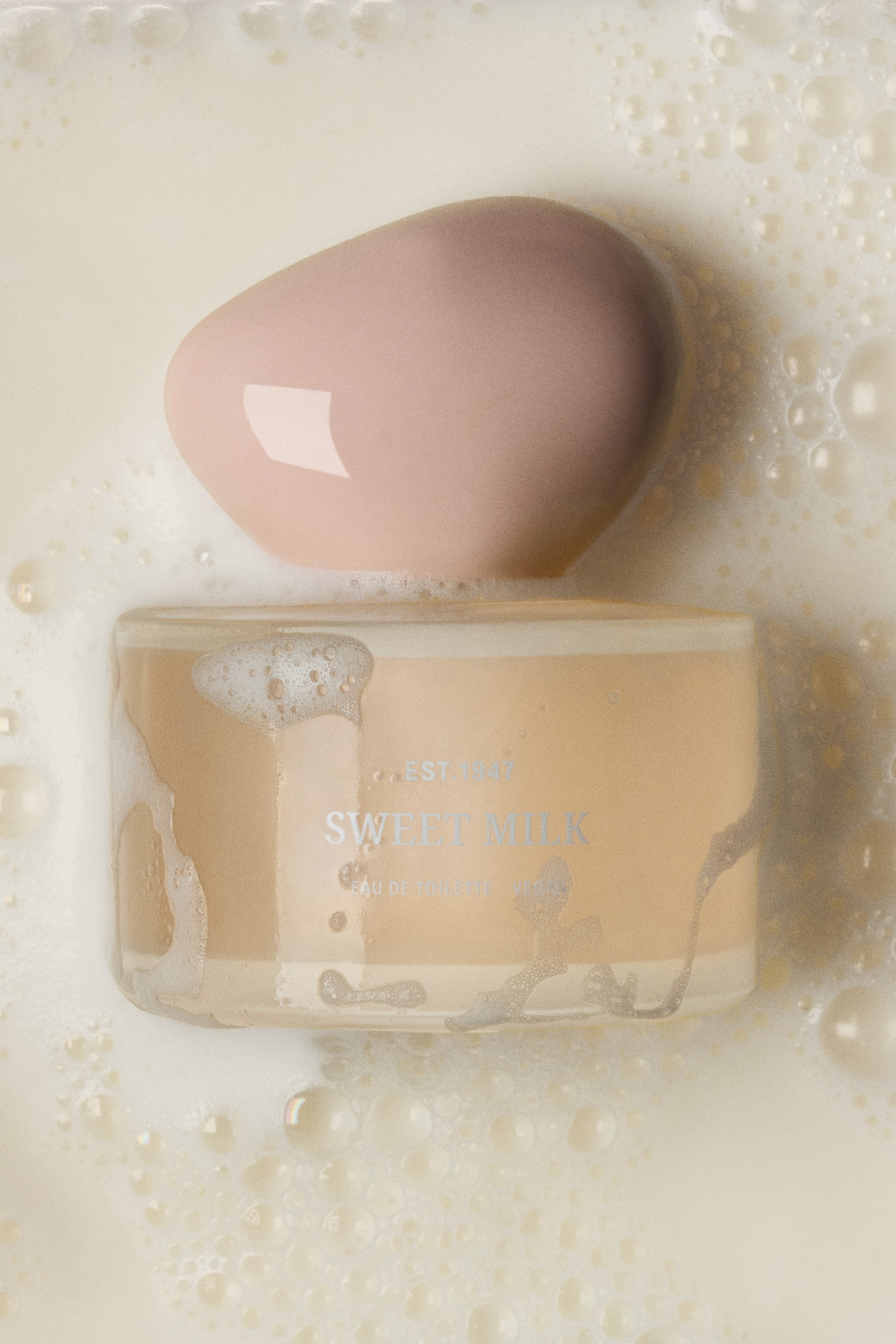 Ver imagem maior: O frasco de Eau De Toilette Sweet Milk é apresentado numa perspetiva de cima, parcialmente submerso num líquido cremoso, de tom creme, com espuma, enquanto bolhas aderem à sua superfície de vidro. O frasco cilíndrico contém um líquido bege-claro e é rematado por uma tampa lisa, fosca, de tom rosa, em forma de seixo.
