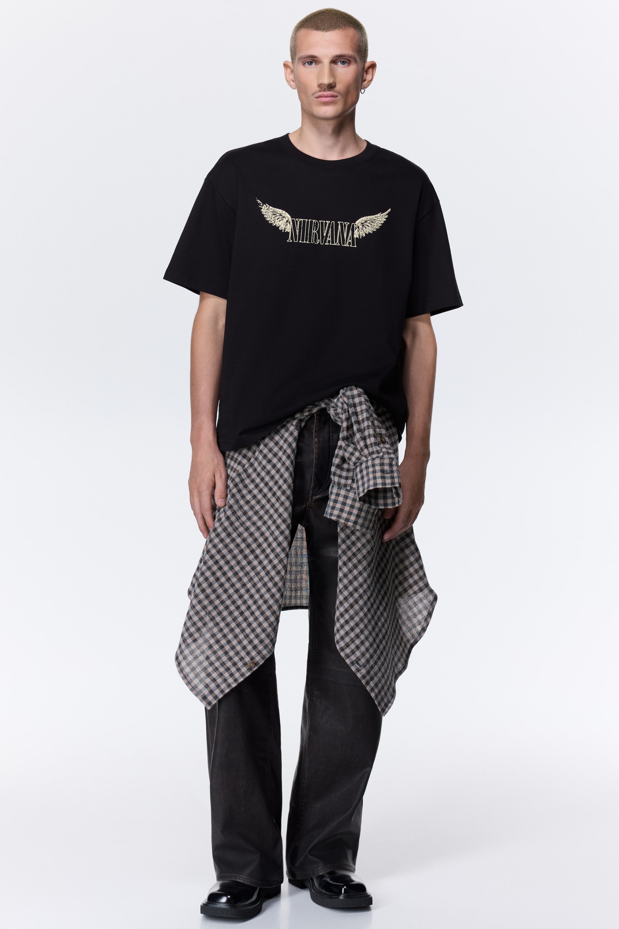 Xem ảnh lớn hơn: Mẫu đang diện một chiếc áo thun màu đen in đồ họa “Nirvana” kèm đôi cánh. Áo có cổ tròn bo gân, vai xuôi và gấu áo cắt thẳng, phom rộng rãi, được phối cùng một chiếc áo sơ mi kẻ caro buộc quanh eo.