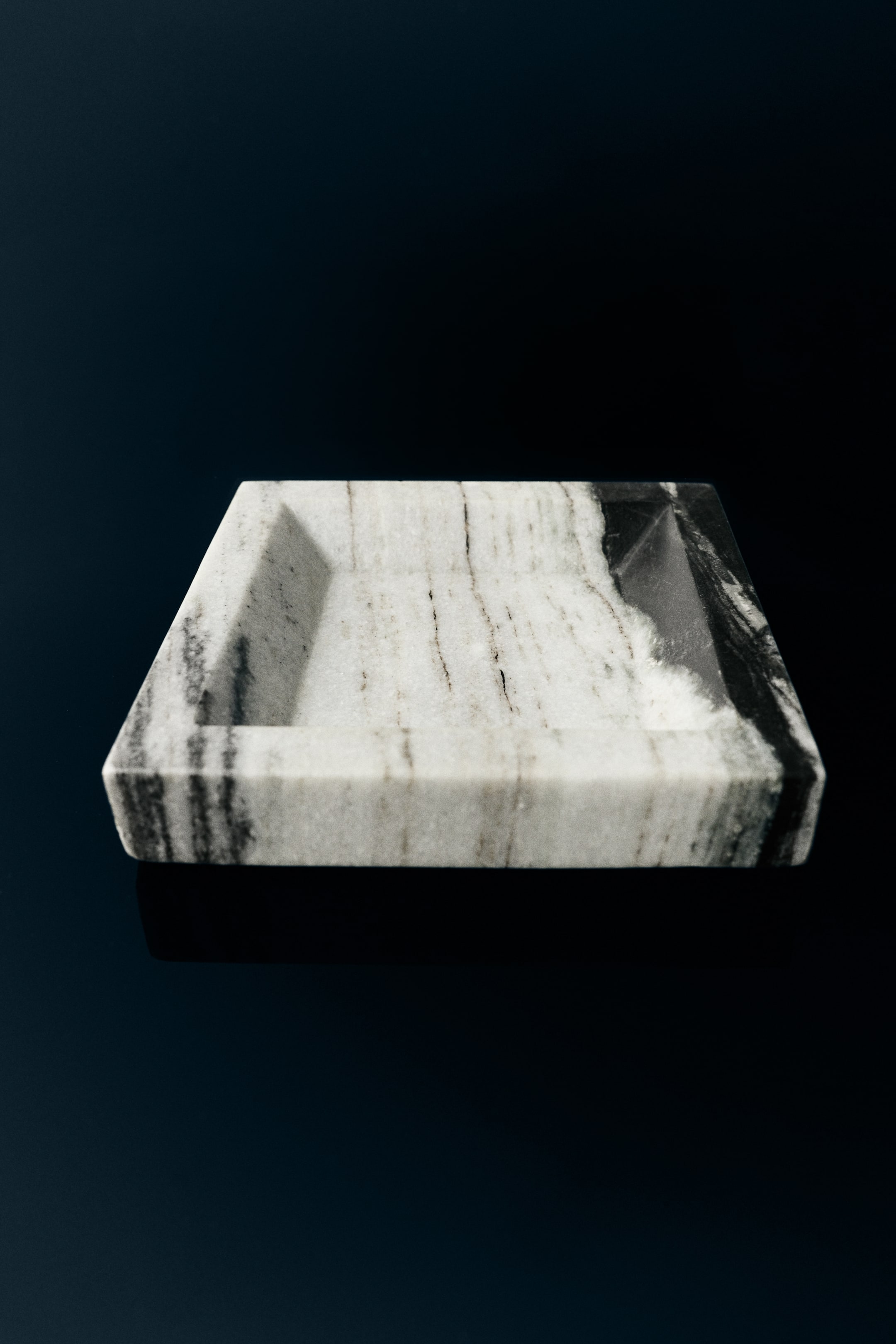 View larger image: A small, square marble tray is positioned at a slightly elevated front angle, highlighting a thick outer rim and sloped interior walls. The tray features distinctive natural veining, with dark grey and black lines running vertically across the white marble surface.