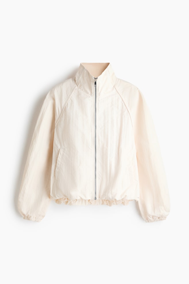 A light beige jacket is presenting from the front, featuring a stand collar and a full-length silver zipper. It is displaying raglan sleeves, slanted front pockets, and an elasticated hem and cuffs. - 1