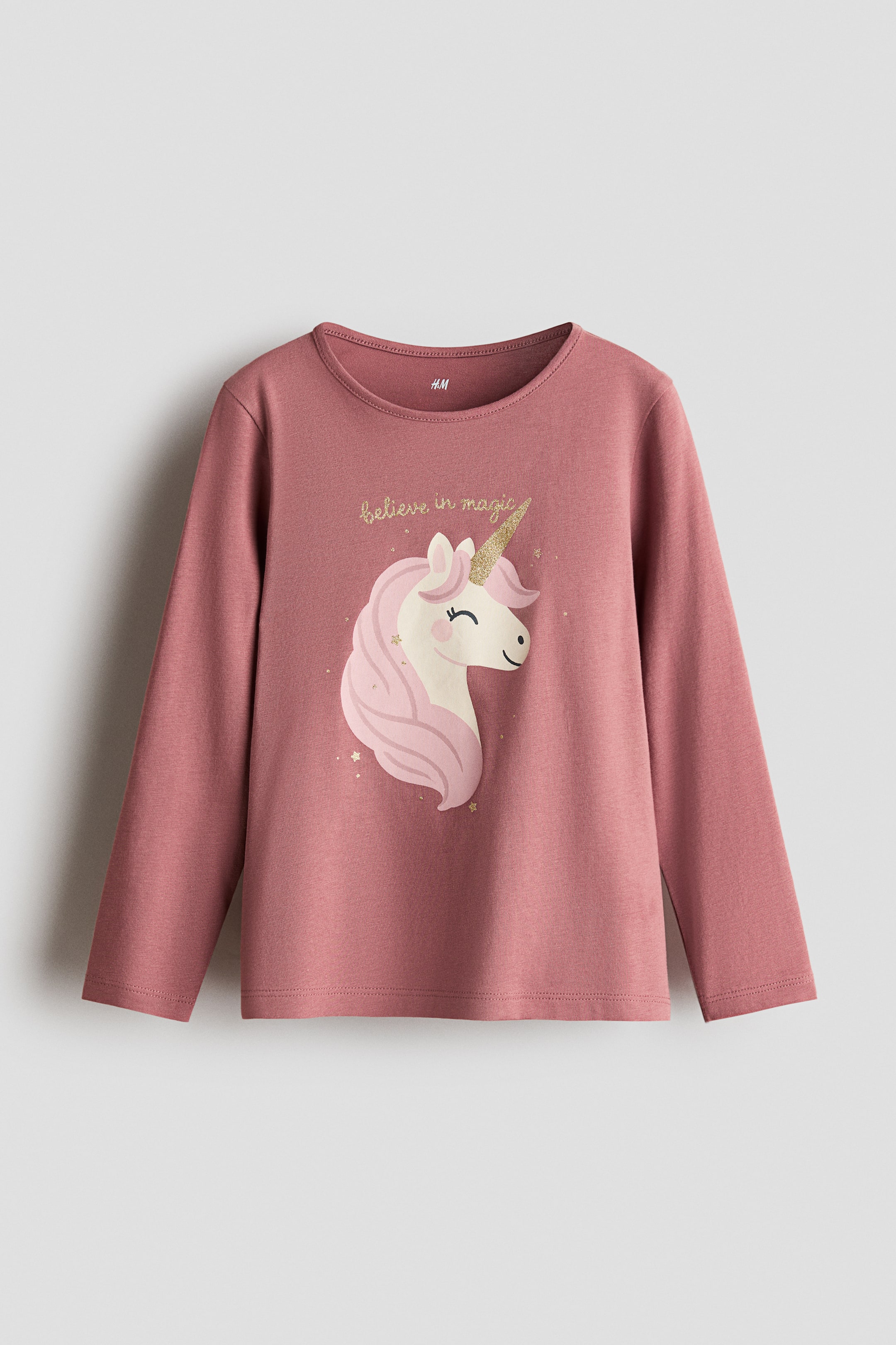 View larger image: A dusty rose long-sleeve top is shown from the front, featuring a round neckline with a narrow trim. The front displays a graphic print of a unicorn with a pink mane and a glittery gold horn beneath the gold script text "believe in magic."