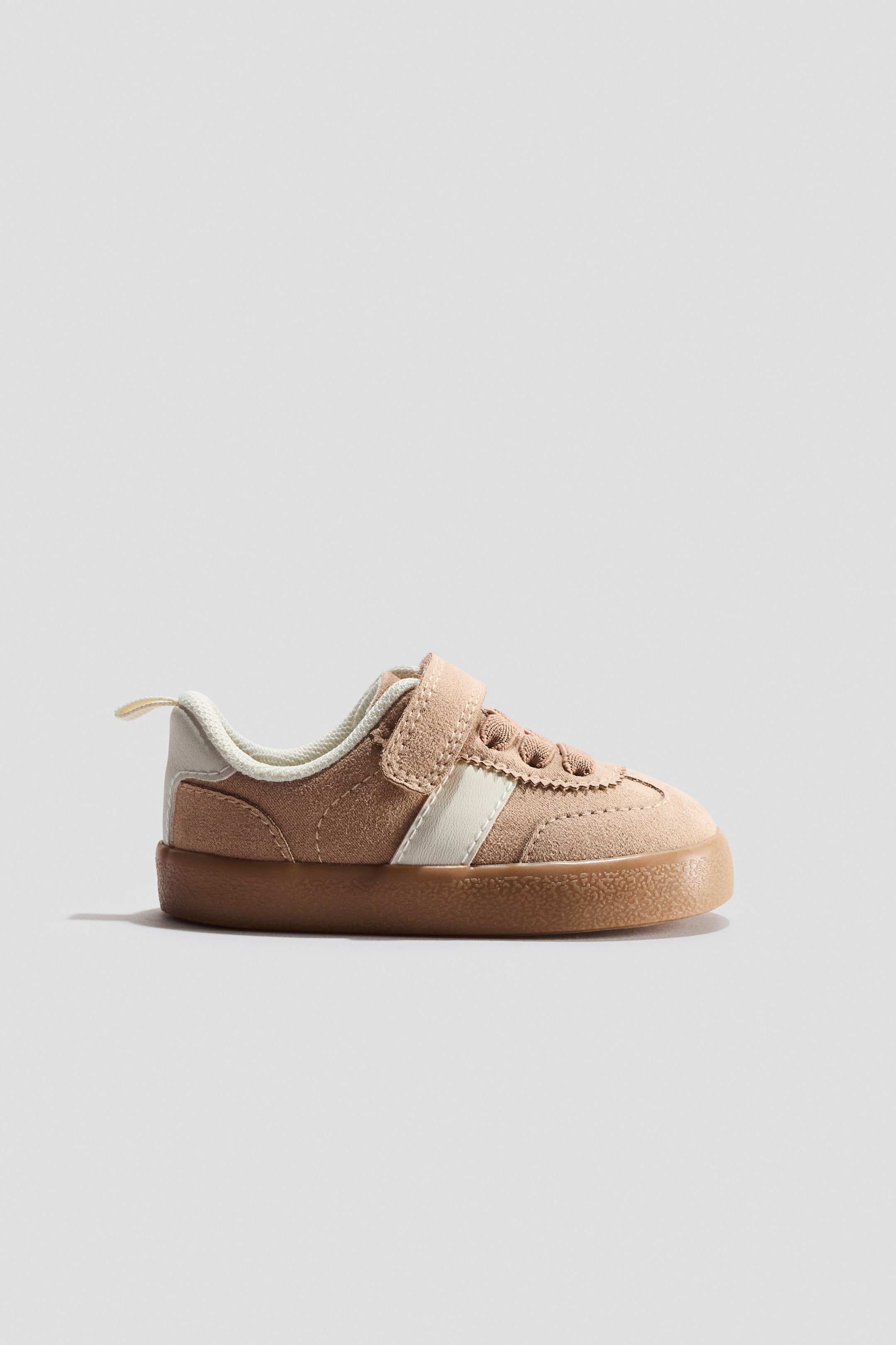 Agrandir l'image: Une basket à profil bas présente une tige de couleur marron clair semblable à du daim, avec une patte à scratch crème à l'avant, un laçage élastiqué et une languette rembourrée. Elle est agrémentée d'un passepoil crème autour du col, d'une bande crème sur le côté, d'un contrefort crème et d'une boucle en gros-grain crème à l'arrière, reposant sur une semelle marron à motifs.