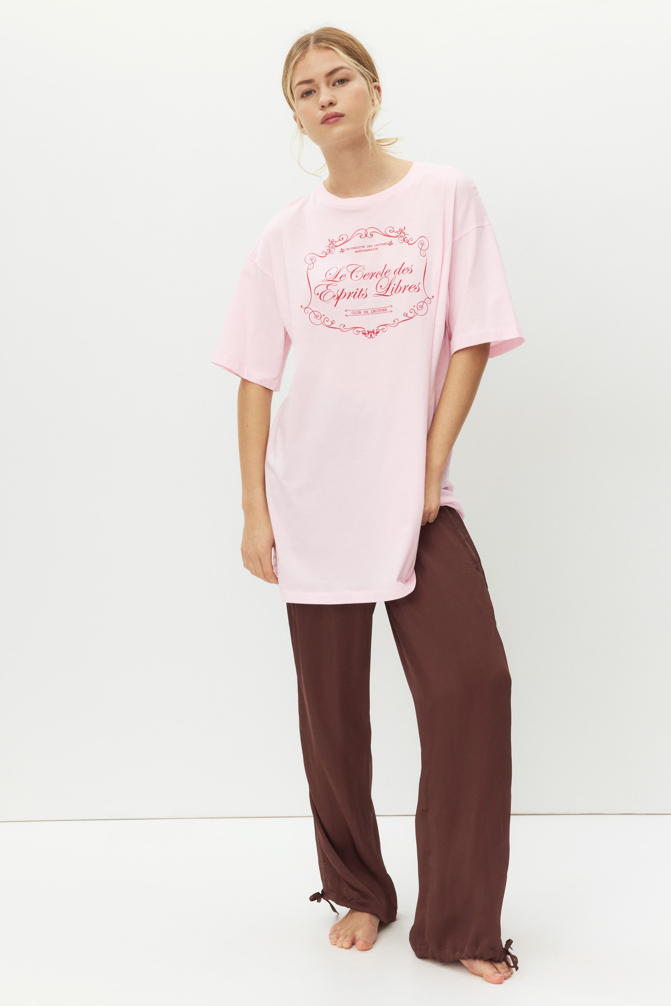 Näytä suurempi kuva: Vaaleanpunainen oversize t-paita esitellään, ja sen etupuolella on punainen "Le Cercle des Esprits Libres" -grafiikka koristeellisilla reunoilla. Tummanruskeat leveälahkeiset housut ovat käytössä, ja ne esittelevät nilkoissa kangasta kiristäviä nauhoja.