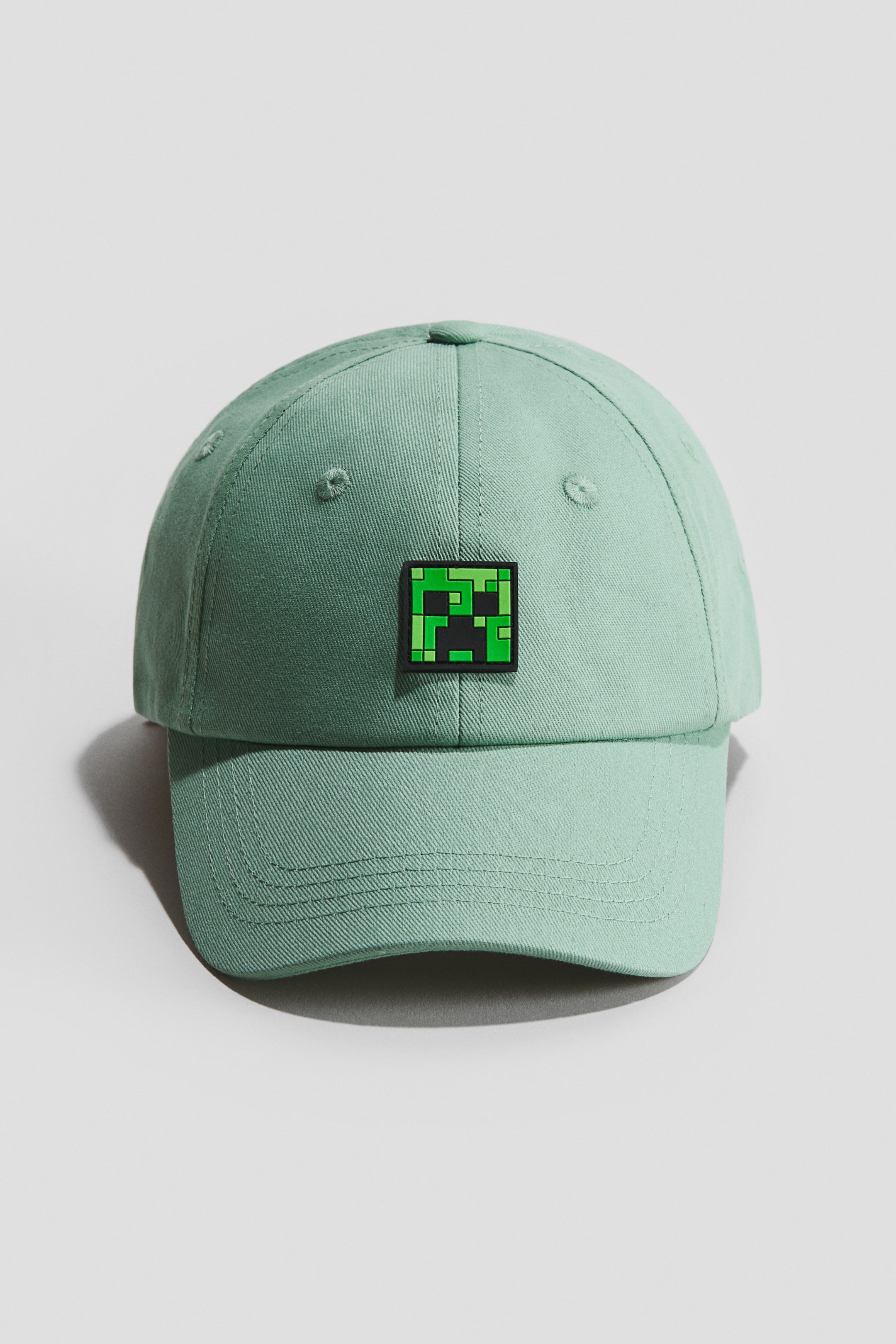הצגת תמונה גדולה יותר: כובע בצבע ירוק בהיר עשוי טוויל כותנה מוצג כ-FRONT, עם מוטיב פנים מרובע מפוקסל בירוק ושחור על הפאנל הקדמי. הכובע מציג גם חורי רקמה ושוליים תפורים.