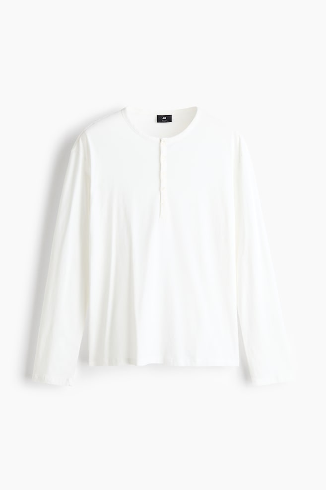 Ein langÃ¤rmliges, cremeweiÃes Henley-Shirt ist von vorne zu sehen und verfÃ¼gt Ã¼ber einen Rundhalsausschnitt mit Knopfleiste. Das Shirt zeigt eine lockere Passform mit gerade geschnittenen Ãrmeln und einem schlichten, sauberen Saum. - 1