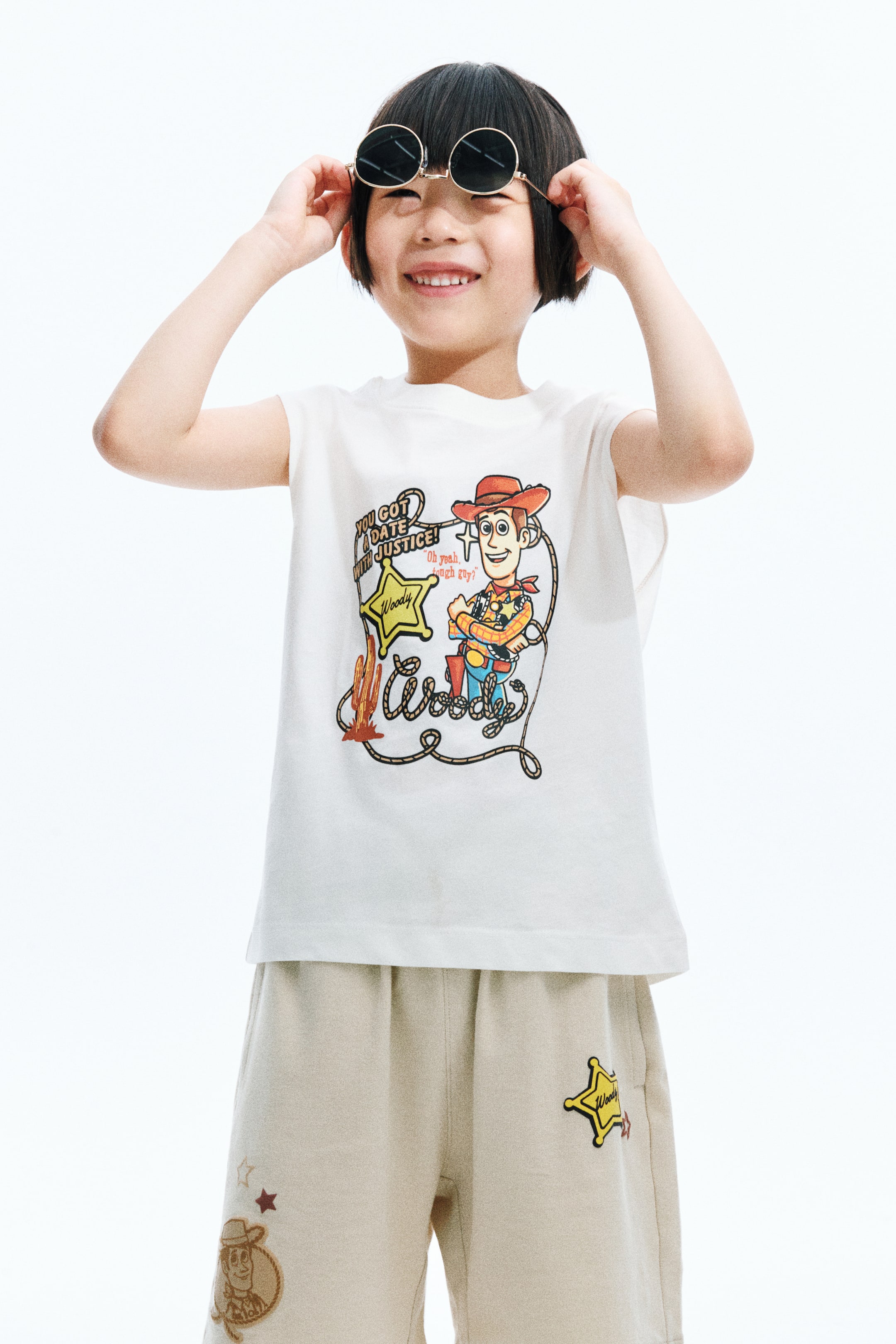 View larger image: A white sleeveless t-shirt paired with matching beige shorts forms a coordinated set. The t-shirt showcases a vibrant graphic of Woody from Toy Story, featuring a yellow sheriff's star with "Woody" and additional text. The shorts display a yellow sheriff's star with "Woody" on the right leg and an outline graphic of Woody's head on the left leg.