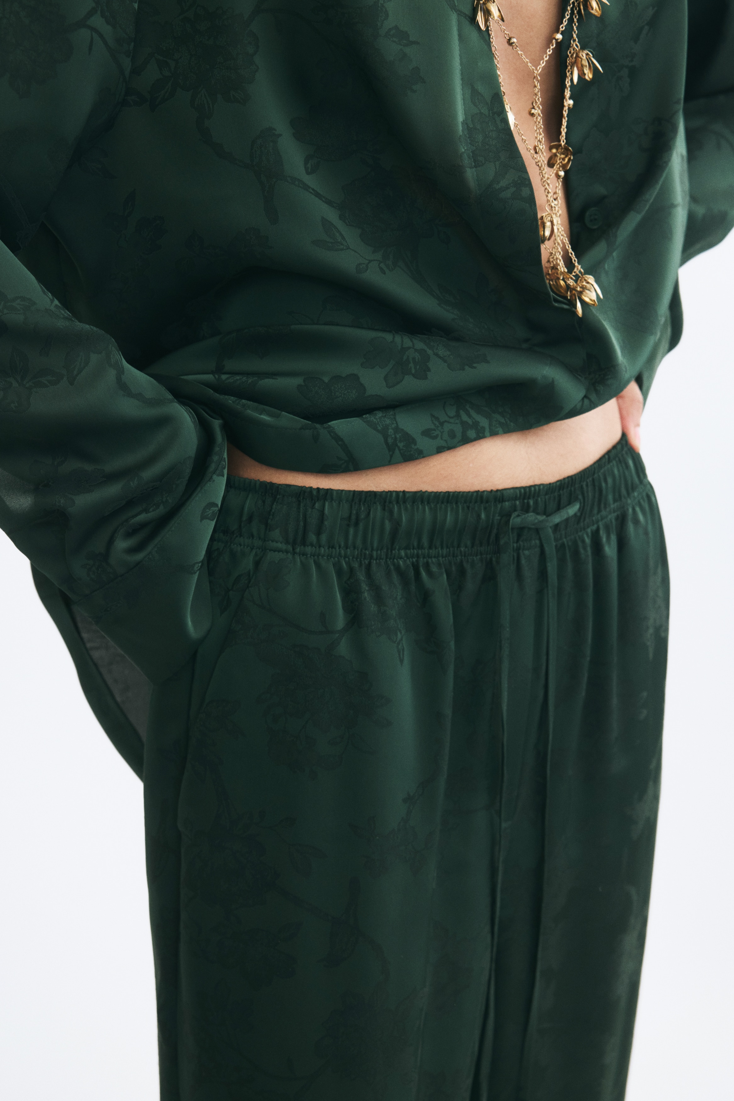 A pair of dark green relaxed-fit trousers in a jacquard weave is hanging front-facing, showcasing a tonal floral pattern and wide, straight legs. The trousers feature an elasticated waistband with a thin drawstring and diagonal side pockets.