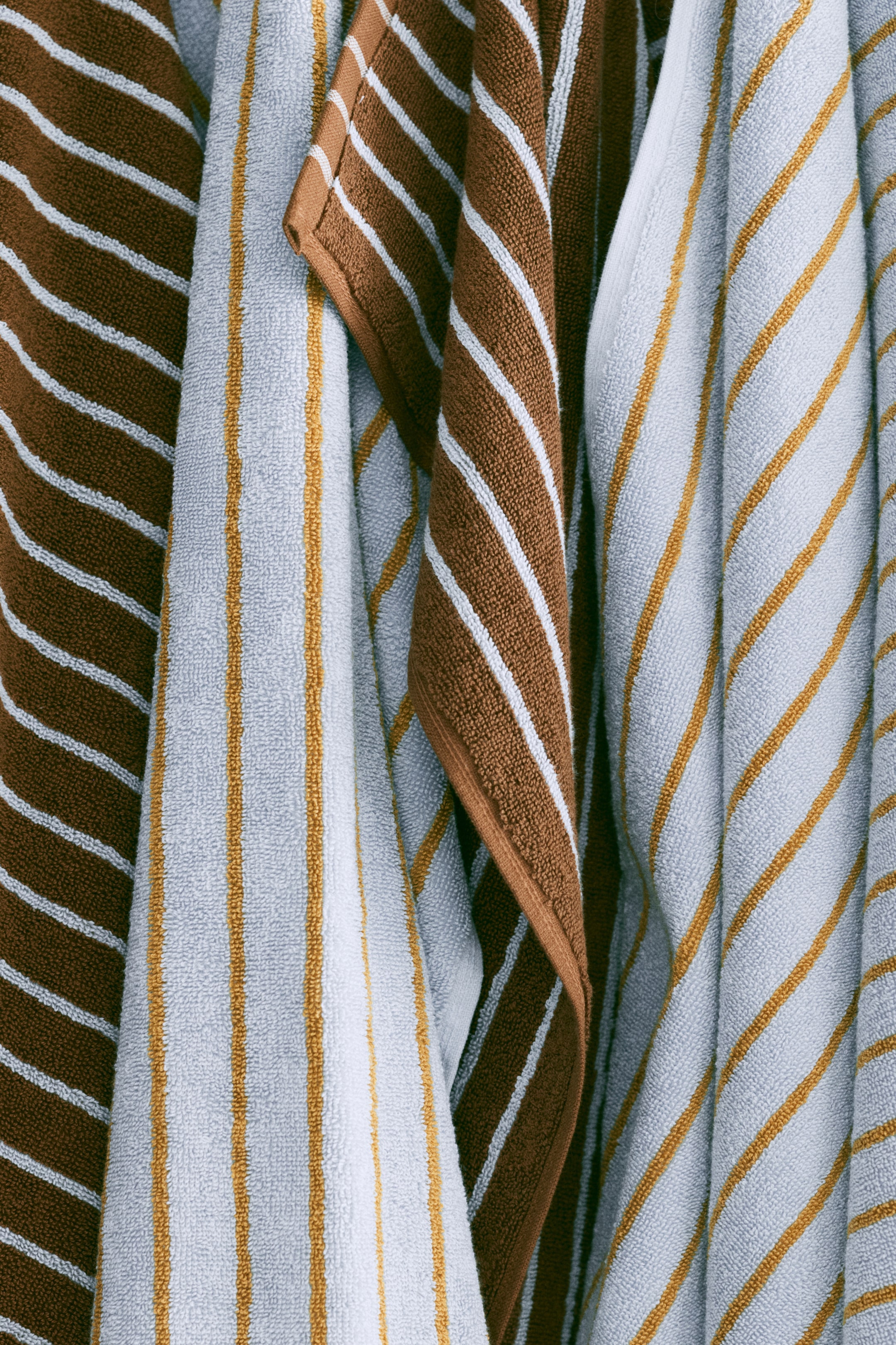 View larger image: Several towels are hung up, showcasing unique striped patterns and textured finishes. Some of the towels boast brown with white diagonal stripes, while others are light blue adorned with golden yellow vertical and diagonal stripes, with a brown towel elegantly draped over a light blue one.