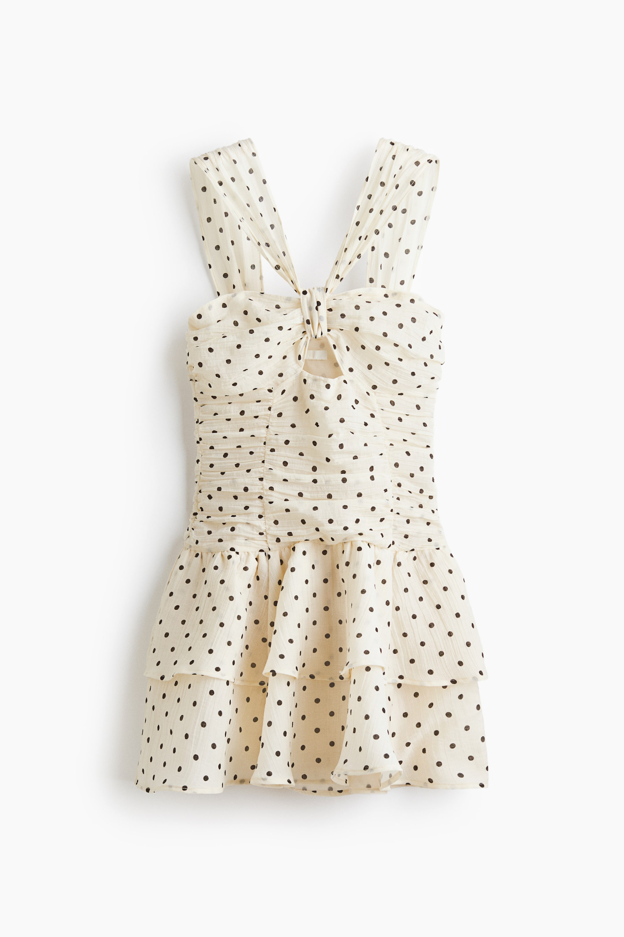 View larger image: A cream mini-dress with black polka dots is displayed front-facing. It features wide shoulder straps that tie into a bow at the bust, revealing a small cutout beneath. The bodice showcases extensive ruching, and the skirt forms three distinct ruffled tiers.