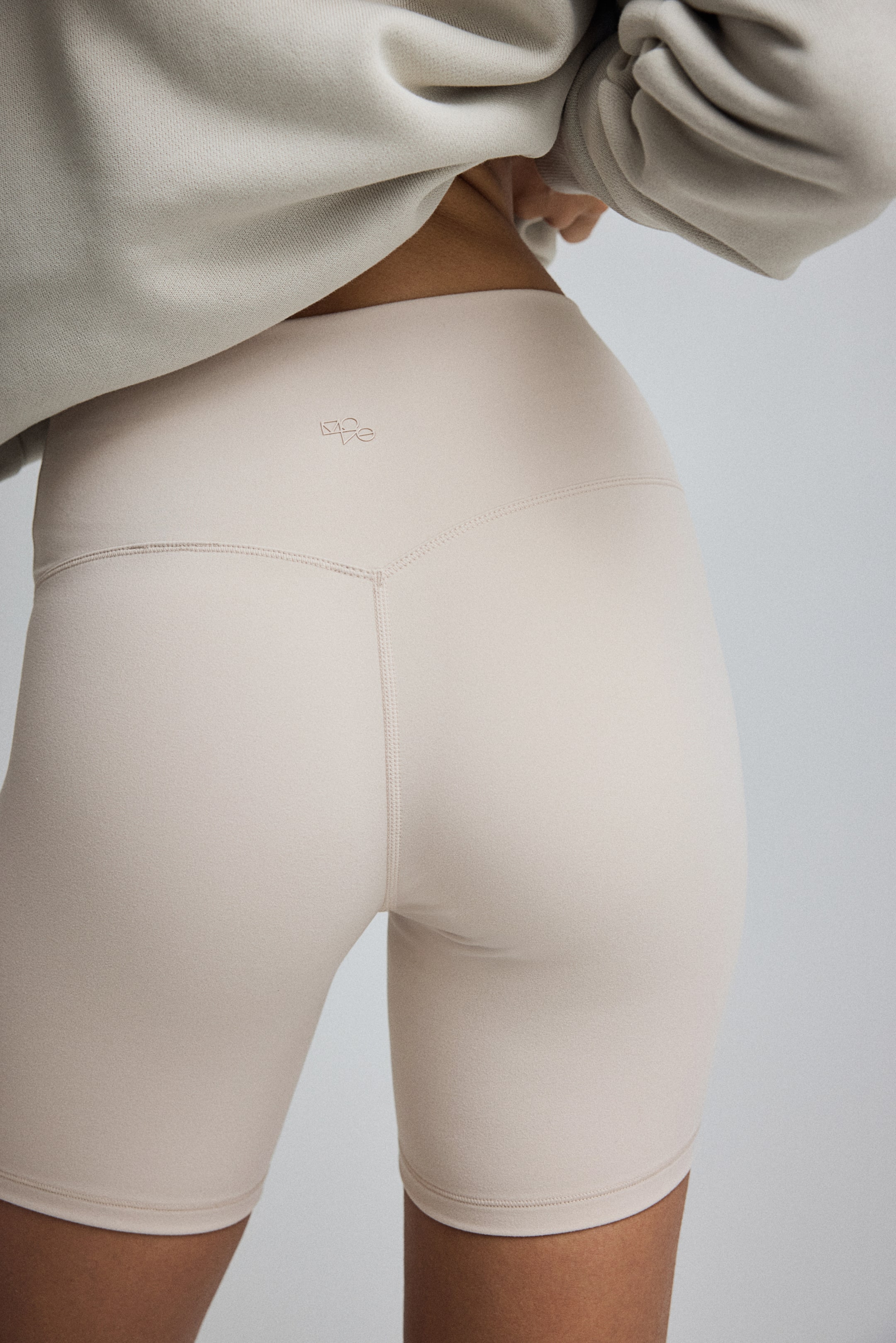 Visualizza immagine più grande: I pantaloncini da biker beige sono presentati da un'angolazione posteriore, aderenti con un design a vita alta caratterizzato da una cucitura a forma di V sulla cintura posteriore e un piccolo logo discreto centrato sopra di essa. I pantaloncini si estendono fino a metà coscia e mostrano cuciture flatlock lungo le gambe.