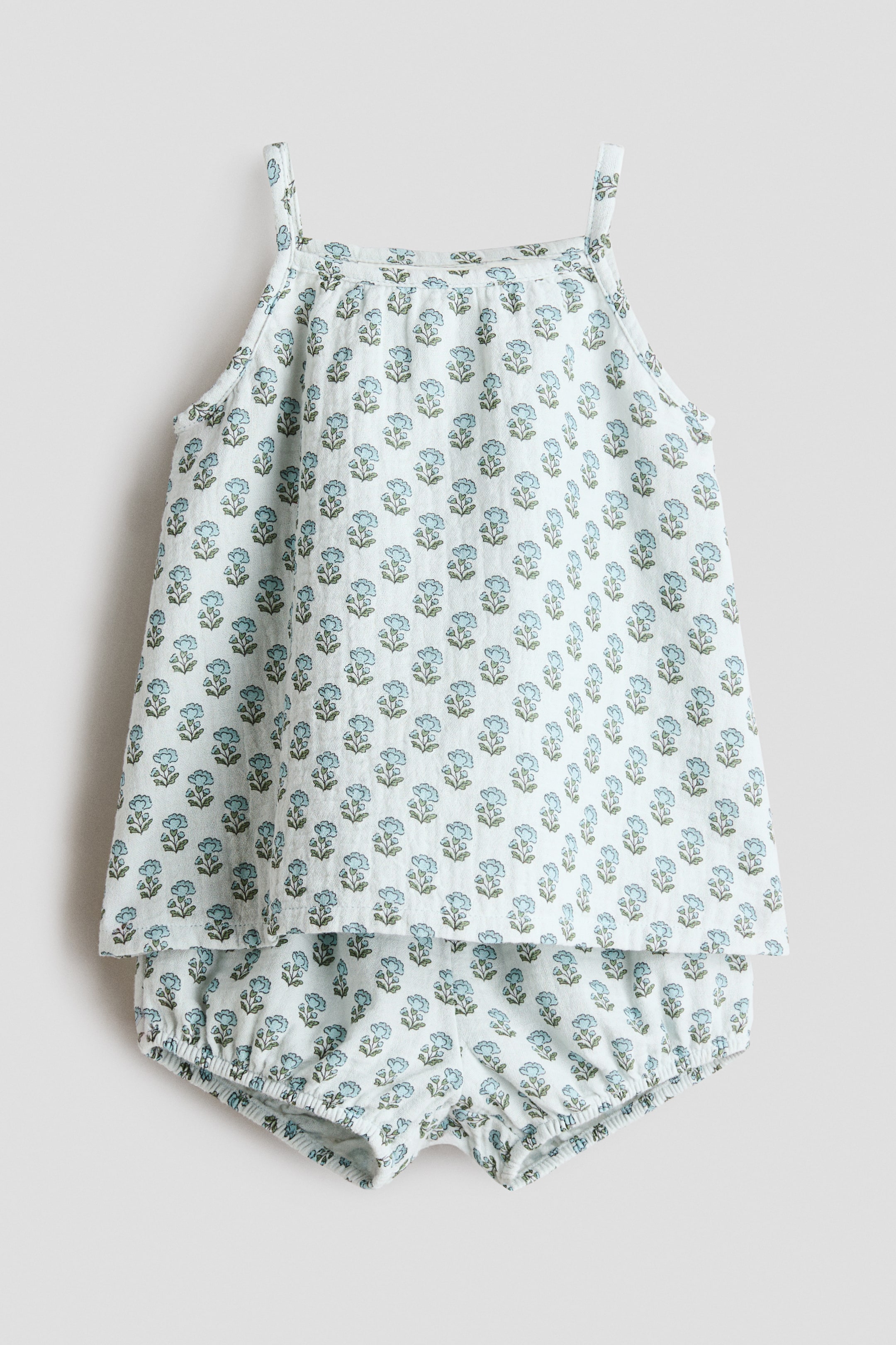 Voir une image plus grande: Un ensemble pour bébé en deux pièces présente un motif floral bleu clair avec des feuilles vertes sur un fond pâle, vu de face. Le haut sans manches est conçu avec des bretelles fines et un col froncé, tandis que les shorts bloomer assortis montrent des ouvertures de jambes élastiquées.
