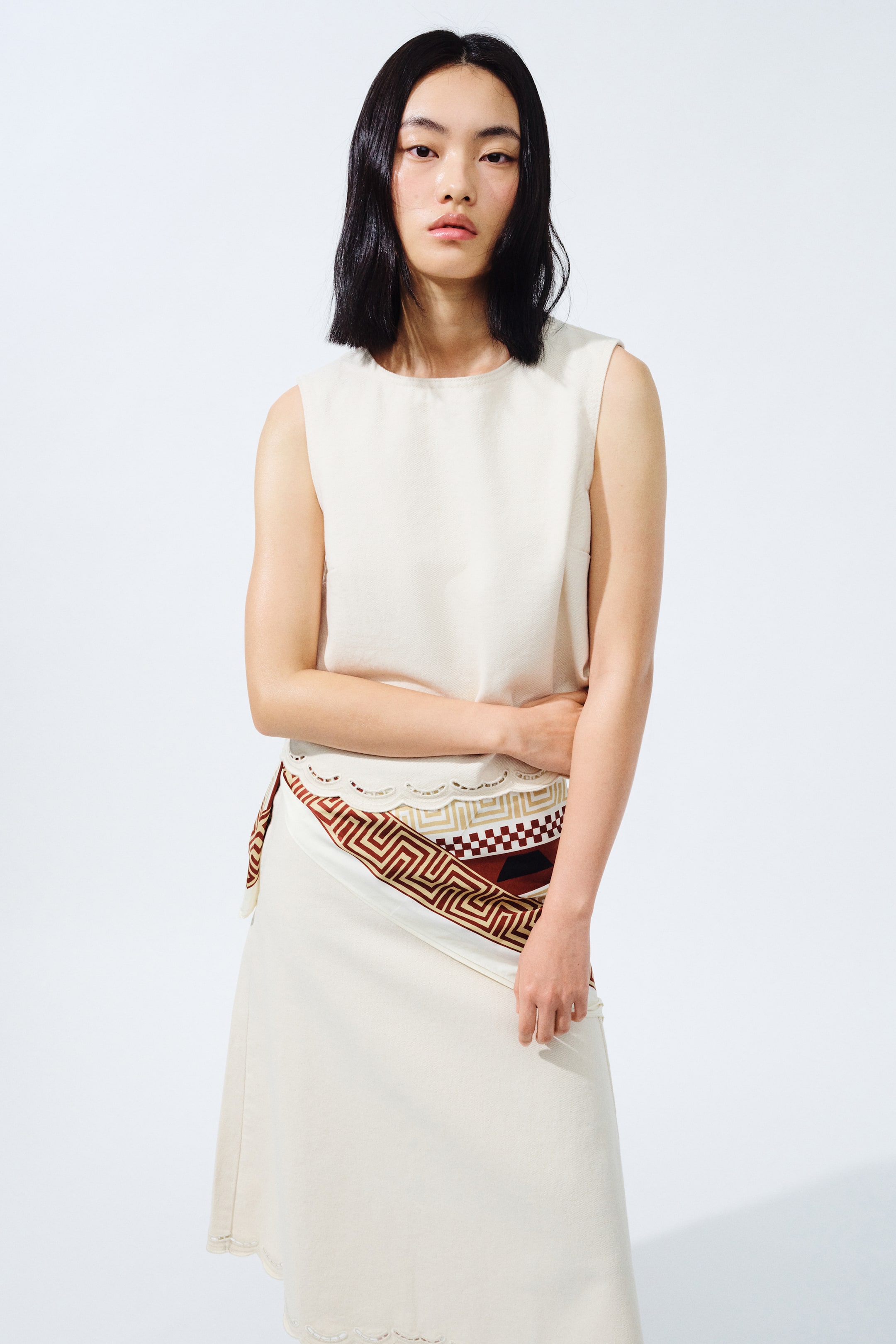 View larger image: A sleeveless top berwarna putih pudar dengan leher bulat sedang dipakai. Skirt midi berwarna putih pudar, dengan perincian potongan scallop pada bahagian bawah dan lapisan atas, digayakan bersama sash bercorak yang memaparkan reka bentuk geometri dalam warna coklat, merah, dan putih pudar yang dililit di sekeliling pinggang.