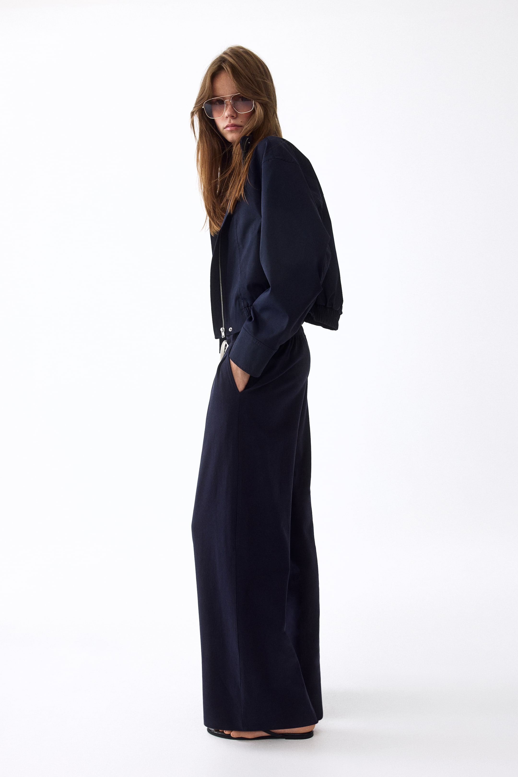 View larger image: Presenting a navy blue two-piece set, consisting of a cropped bomber jacket with a partially unzipped front zipper and ribbed cuffs. Complementing it are wide-leg trousers featuring an elasticated waistband and side pockets, with one pocket casually holding a hand.