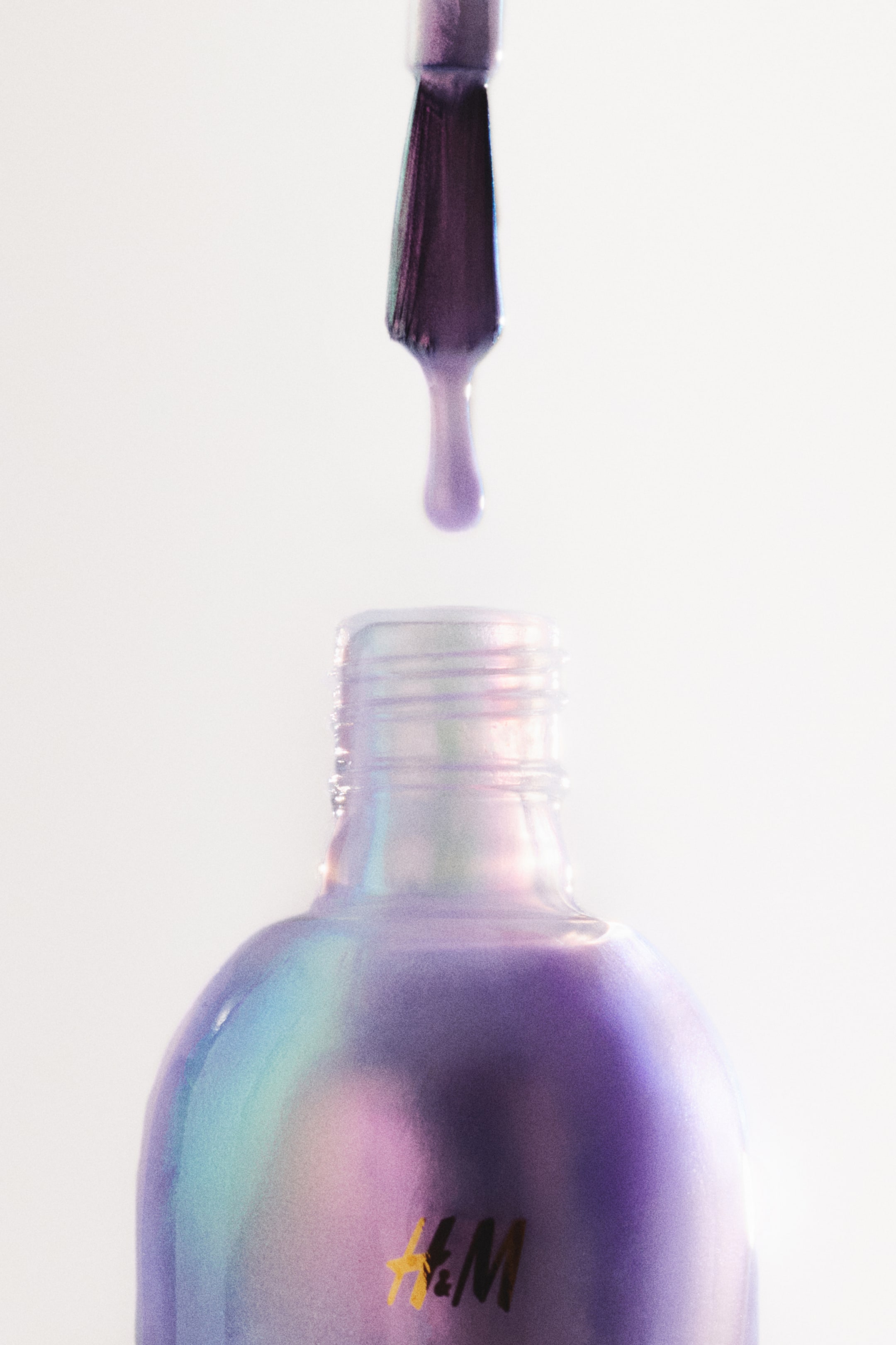 Visualizza l'immagine ingrandita: Una bottiglia di smalto per unghie olografico viola mostra un logo dorato H&M. Un pennello per smalto è sospeso sopra l'apertura della bottiglia, con una goccia di smalto viola che cade dal pennello.