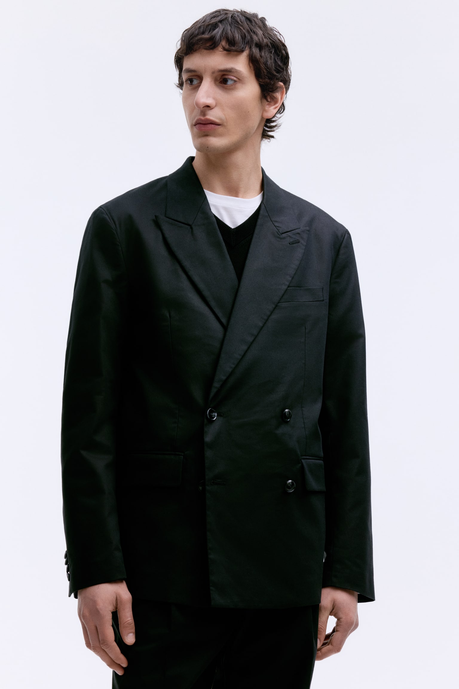 The model is wearing a black double-breasted blazer with a relaxed fit. It includes peak lapels with a buttonhole, a chest pocket, jetted front pockets with flaps, and decorative buttons on the cuffs. - 1