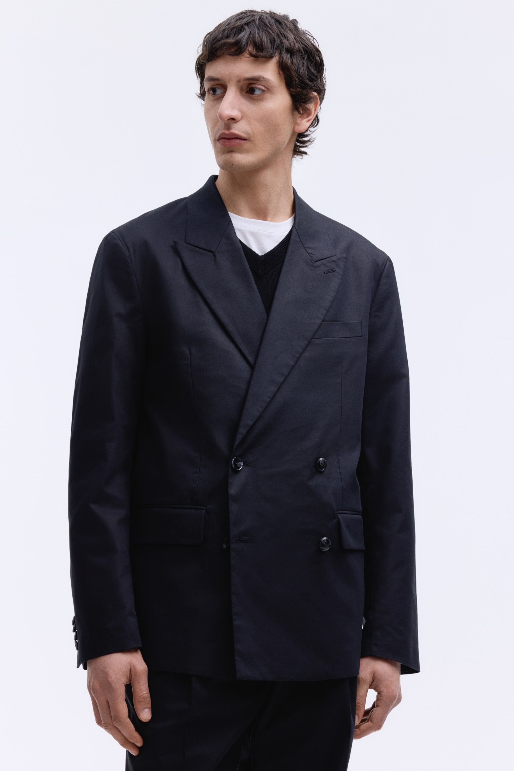 View larger image: The model is wearing a black double-breasted blazer with a relaxed fit. It showcases peak lapels with a buttonhole, a chest pocket, jetted front pockets with flaps, and decorative buttons on the cuffs.
