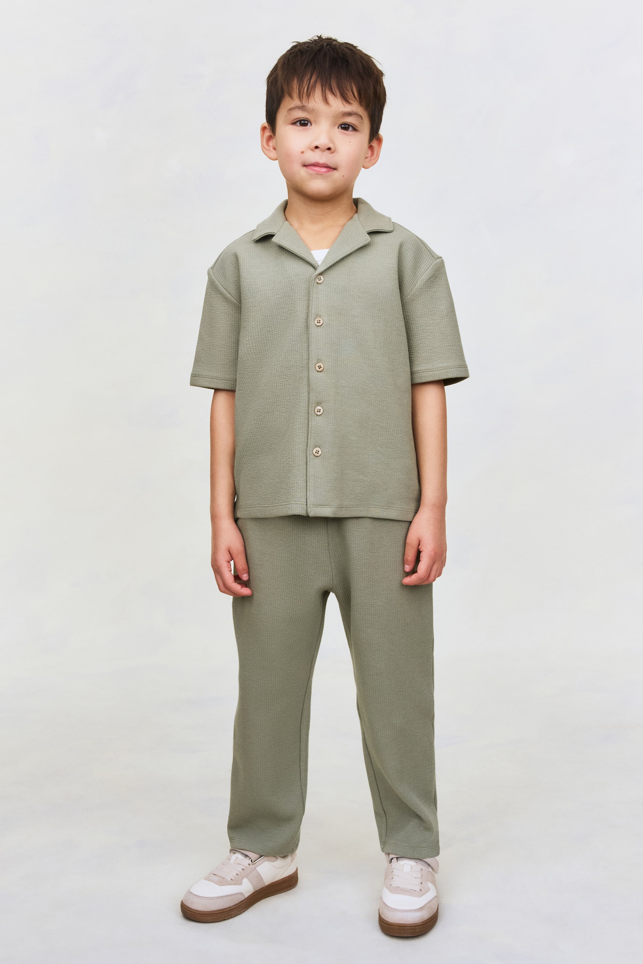 Ampliar la imagen: Un conjunto de dos piezas en verde oliva claro, en tejido de punto texturizado, se muestra de frente y presenta una camisa de manga corta y pantalones largos a juego. La camisa está diseñada con cuello cubano, hombros caídos y una abotonadura frontal de cinco botones, y se lleva sobre una camiseta blanca, mientras que los pantalones de corte relajado complementan la prenda superior.