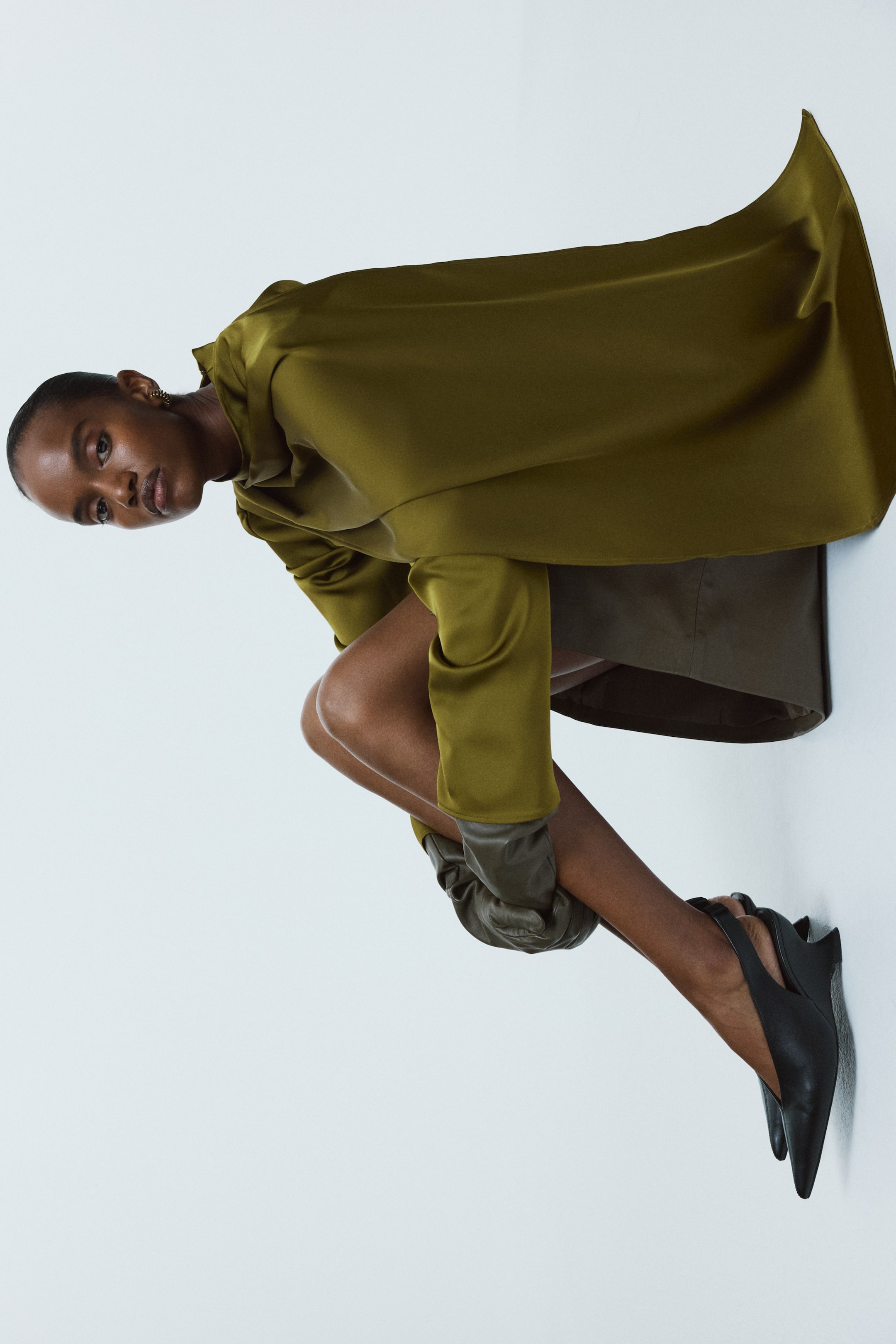 View larger image: An olive-green lustrous satin blouse, featuring a stand collar and a voluminous shawl detail draped across the front, is being worn by the model. Its long sleeves are gathered at the forearms, revealing dark gloves on the model's hands.